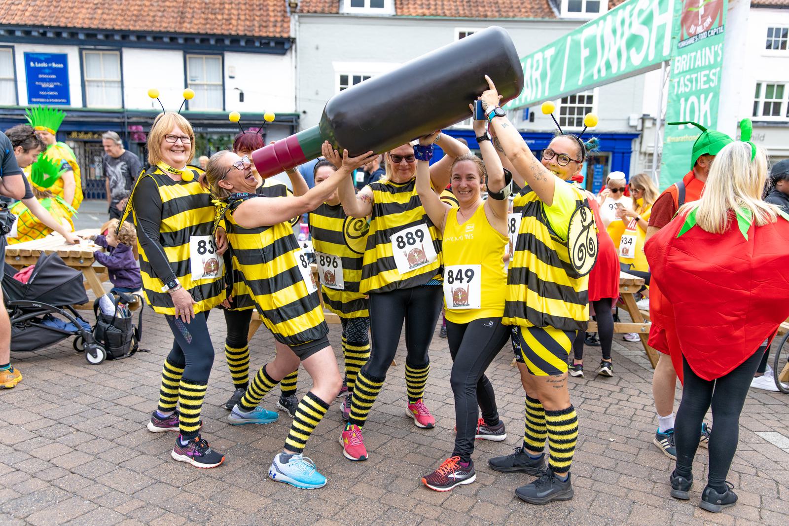 A group of people dressed in bee costumes pose together while lifting a large inflatable wine bottle. They are participating in a fun run event, as indicated by the race bib numbers pinned to their outfits. In the background, other costumed participants and a finish line banner are visible.