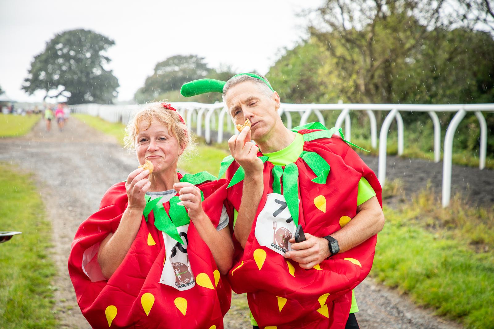 Two adults dressed in strawberry costumes eating snacks during an outdoor event. Both are wearing red, green-topped outfits adorned with yellow seeds and have green hats on. They are standing on a gravel path with fields and trees in the background.