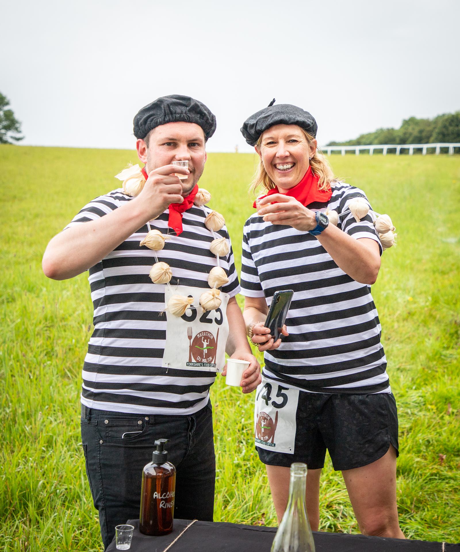 Two people in matching black and white striped shirts, berets, and red scarves stand in a grassy field. One wears a garlic bulb necklace. They are holding up small drink cups and smiling. Both have race numbers pinned on: "45" and "325". A bottle labeled "ALCOOL Riz" is near them.