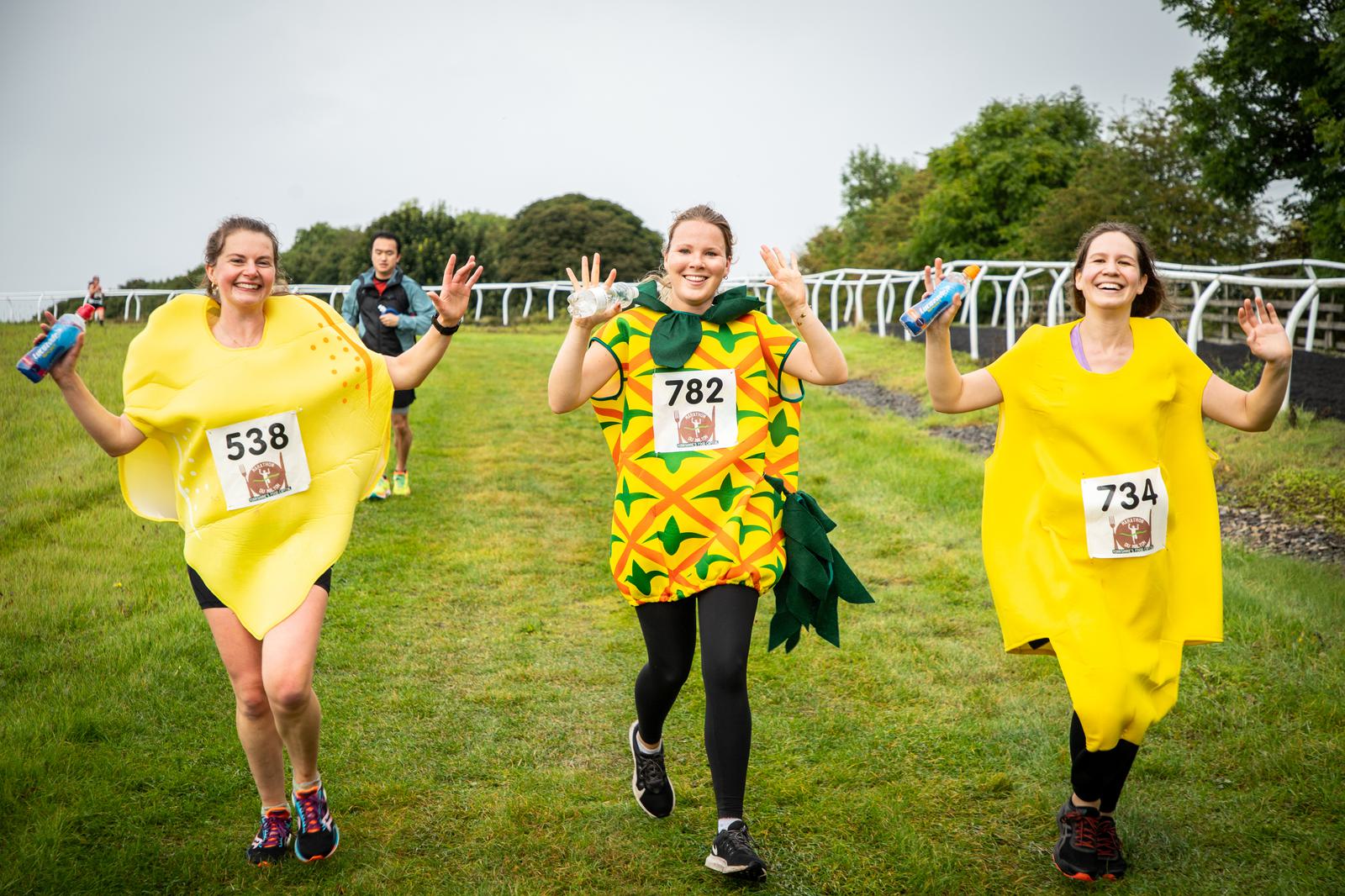 Three smiling runners wearing colorful fruit costumes (a lemon, pineapple, and banana) jog along a grassy path with race numbers 538, 782, and 734. They wave their hands and hold water bottles, with trees and a cloudy sky in the background.
