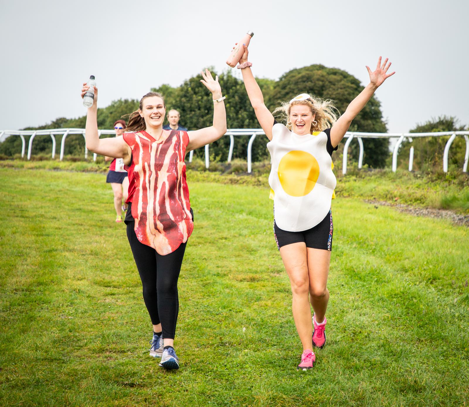 Two people are running on a grassy track, one dressed in a bacon costume and the other in a fried egg costume. Both are smiling and raising their arms in celebration, holding water bottles. Other runners are visible in the background.