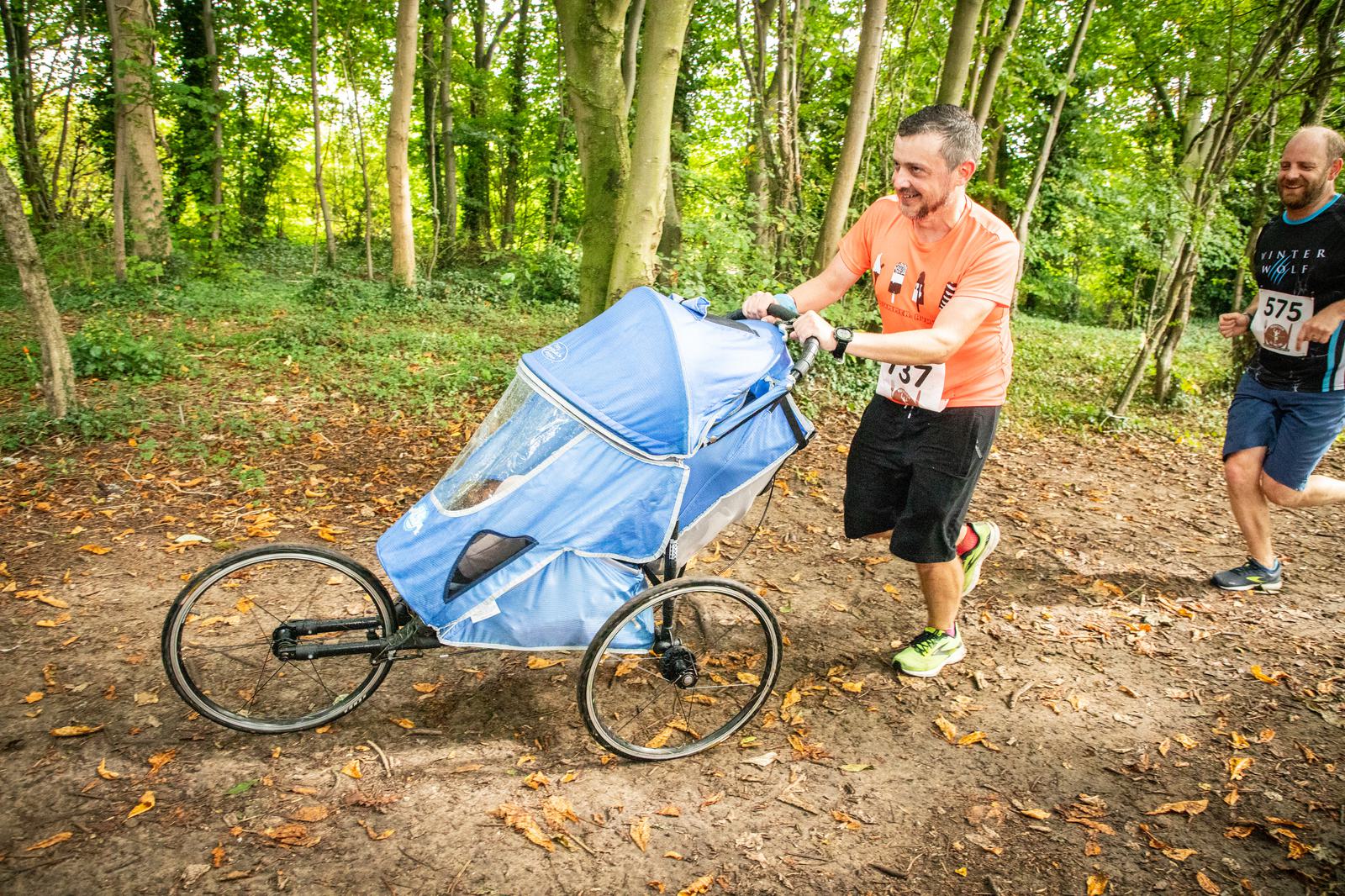 A man in an orange shirt and black shorts is running through a forested path, pushing a blue jogging stroller with a child inside. Another runner in a dark blue shirt and black shorts follows him. Both have race bibs pinned to their shirts.