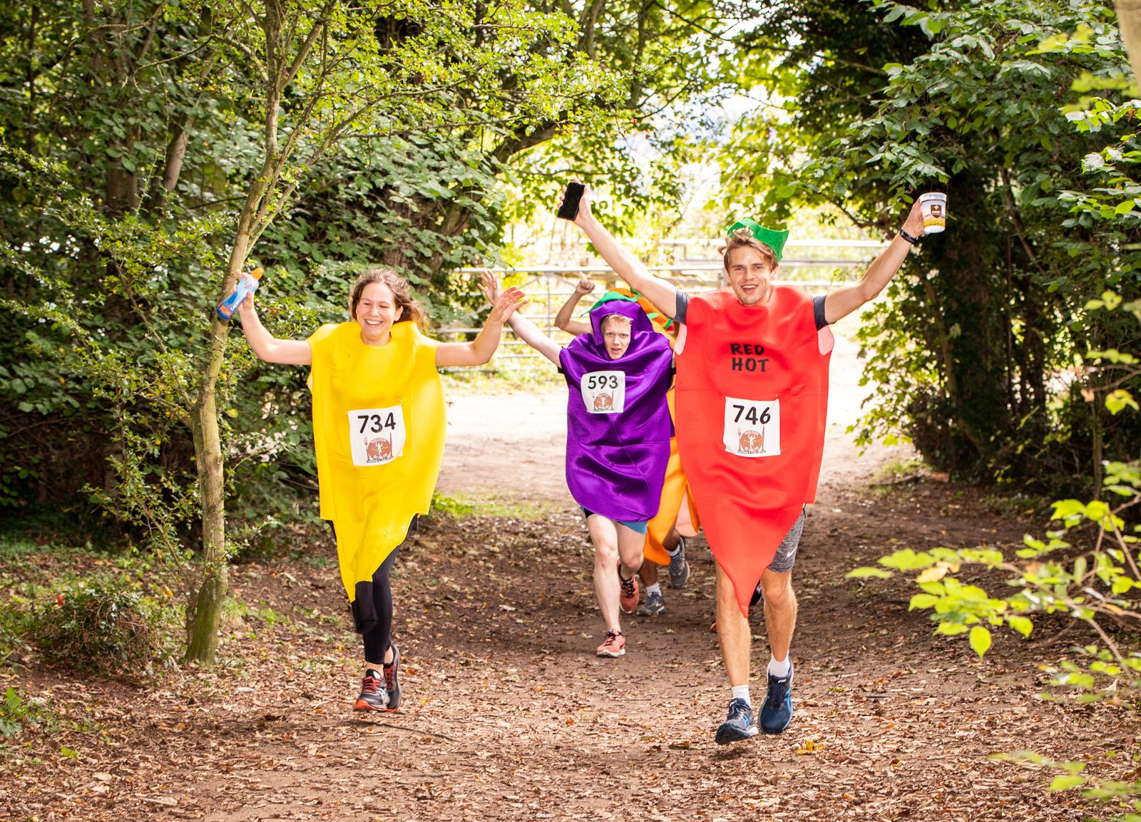 Three people dressed in colorful pepper costumes, yellow, purple, and red, are running in a forested area with their arms raised in celebration. They each wear race numbers (734, 593, and 746) and appear animated and joyful. Trees and foliage surround the path.
