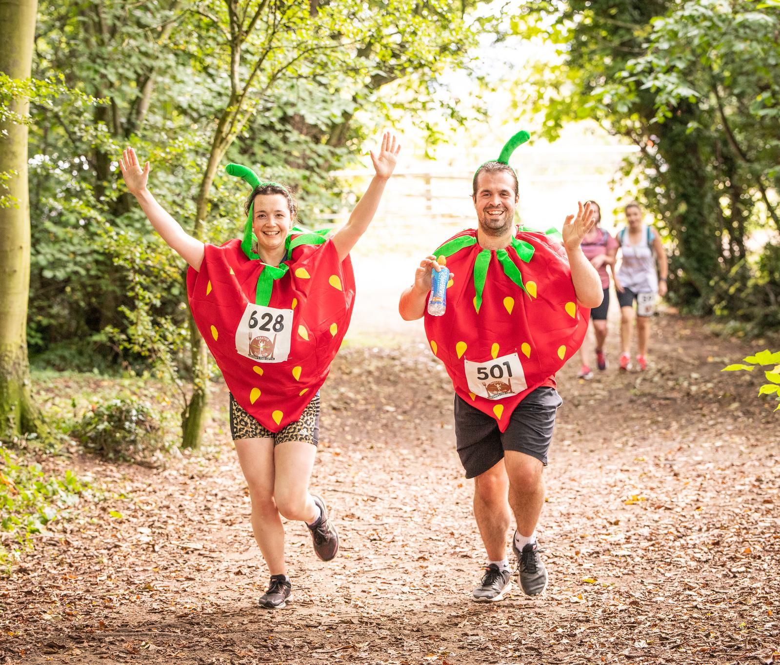 Two smiling people run through a sunlit forest trail dressed in strawberry costumes, both holding their arms up in excitement. They wear race bibs numbered 628 and 501. Other joggers can be seen in the background.