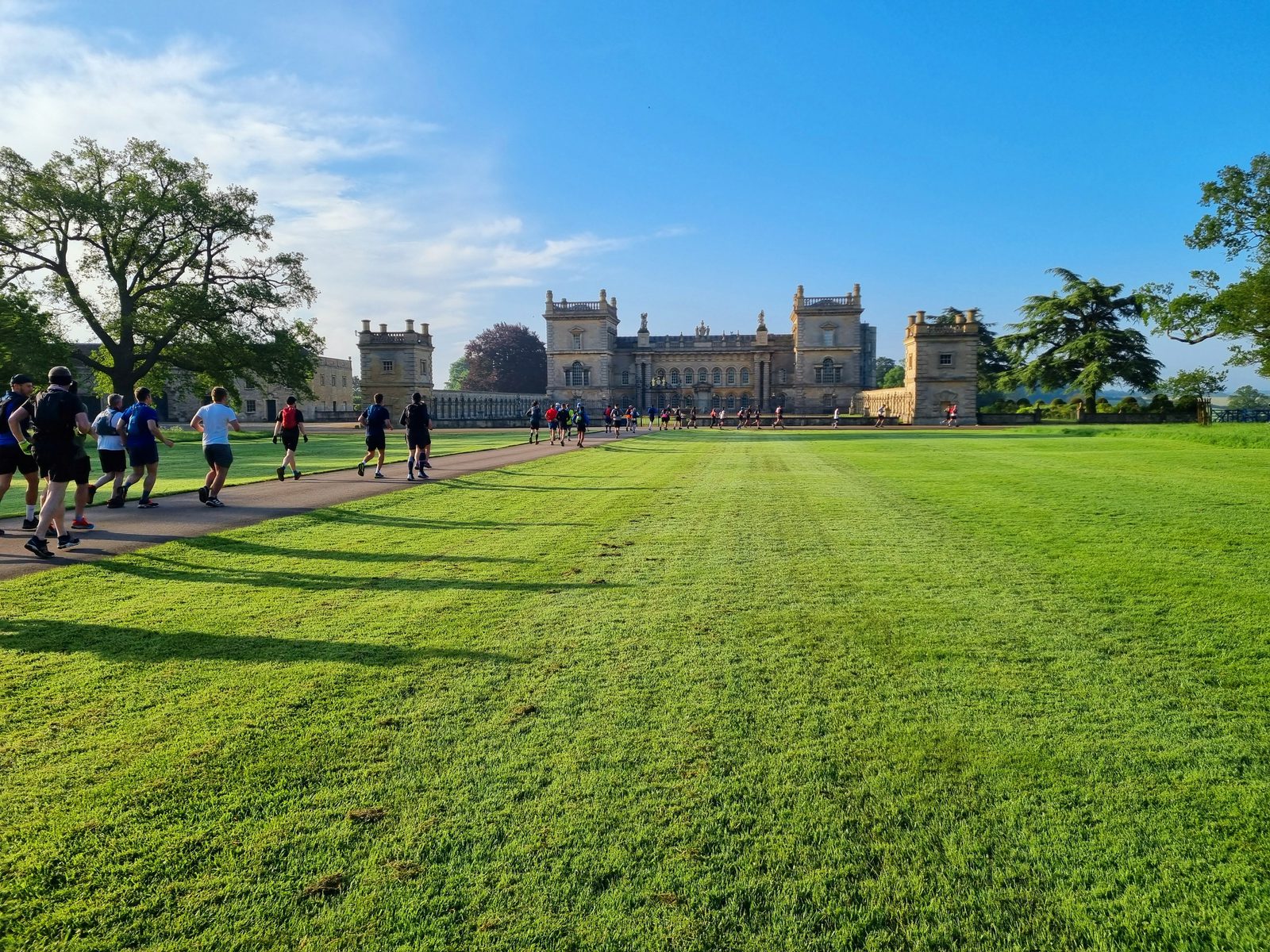 A group of people walking and jogging along a paved path that leads to a grand, historic building with multiple towers. The building is surrounded by lush green lawns and trees under a bright blue sky.