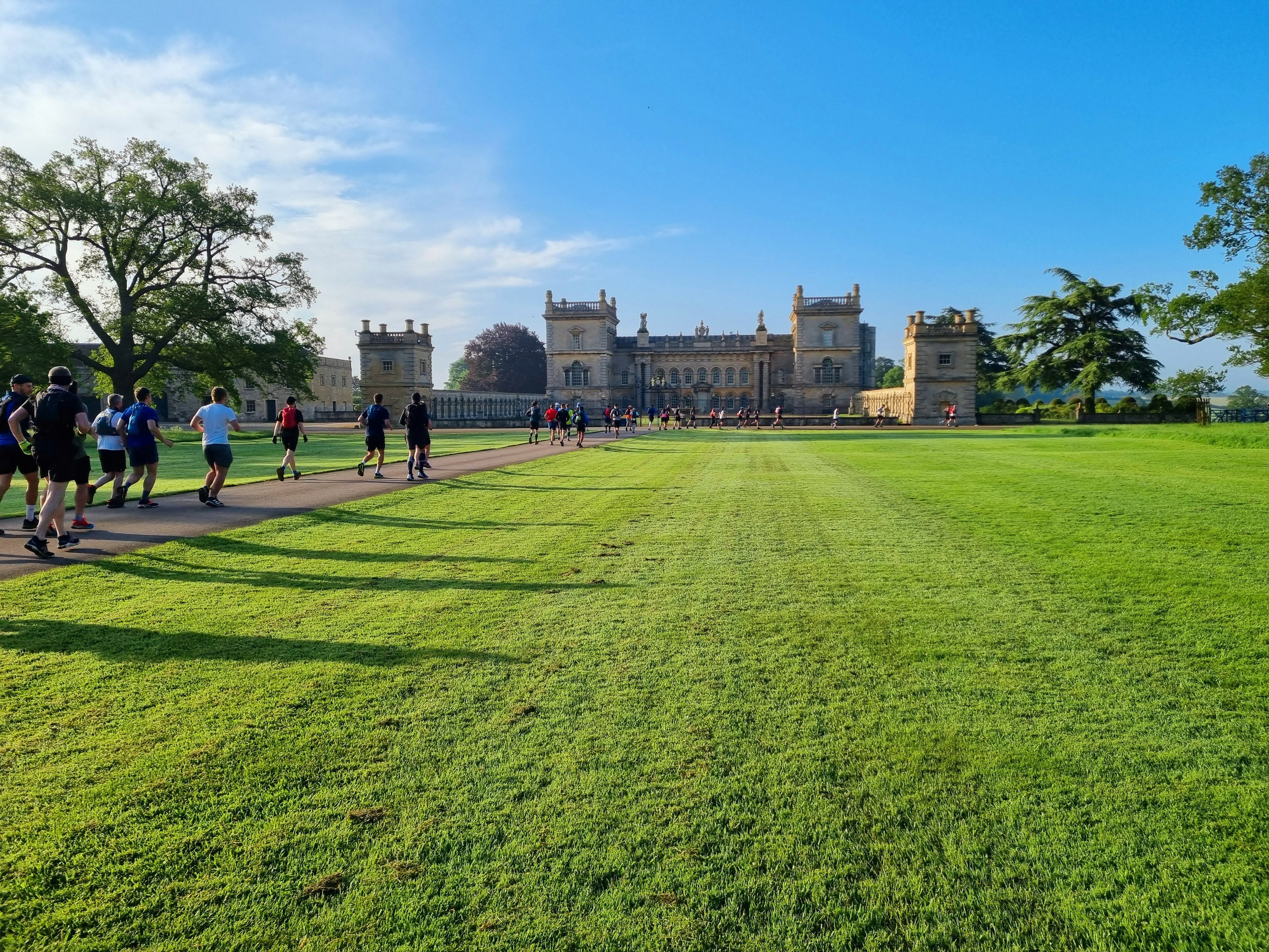 A group of people walking and jogging along a paved path that leads to a grand, historic building with multiple towers. The building is surrounded by lush green lawns and trees under a bright blue sky.