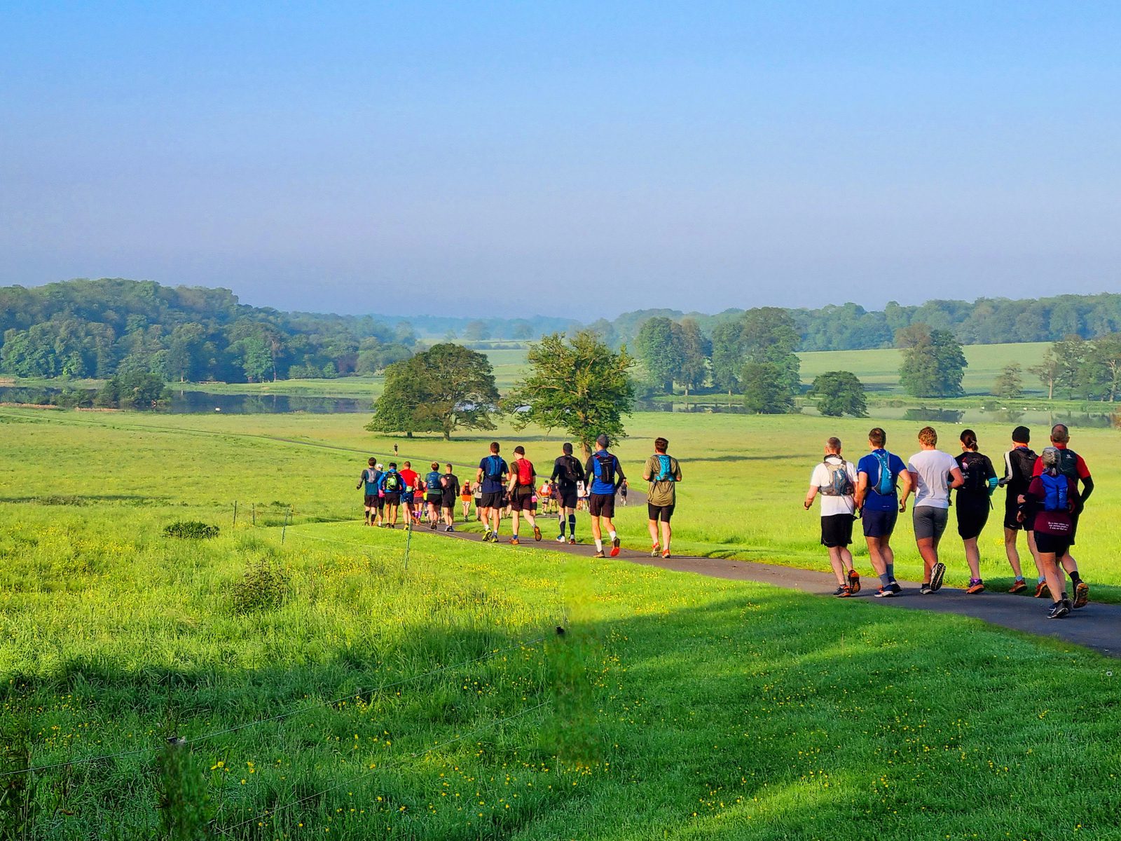 A group of people is running along a paved path through a lush green field. The sky is clear and blue, and trees dot the landscape in the distance. The runners wear various athletic clothes, and some carry backpacks. Sunlight bathes the scene, giving it a vibrant feel.