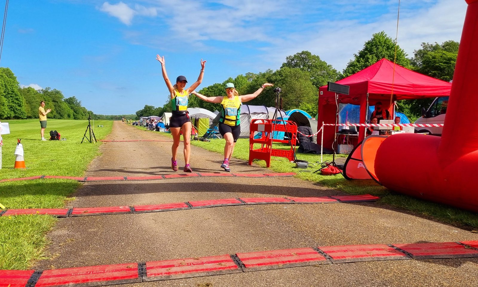 Two runners, wearing yellow tops and shorts, cross the finish line with arms raised in celebration. They are on a paved path lined with red timing mats. Behind them are trees, a row of tents, and a red canopy with people inside and one person cheering. The sky is clear.