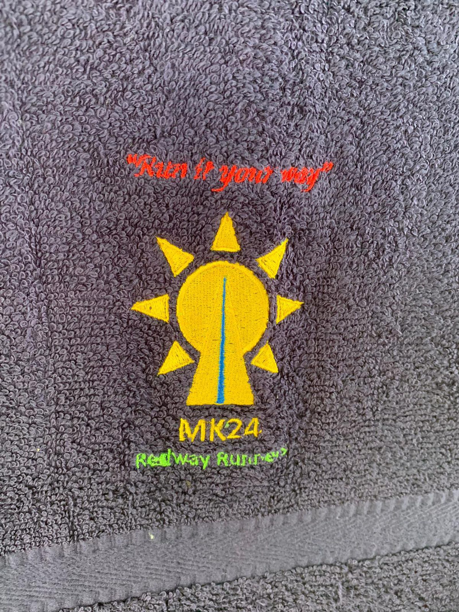 Close-up of a grey fabric with a colorful embroidered logo. The logo features a yellow sun with pointed rays and a blue path in the center. Text above reads "Run it your way," and below it says "MK24 Redway Runners" in red and green text.