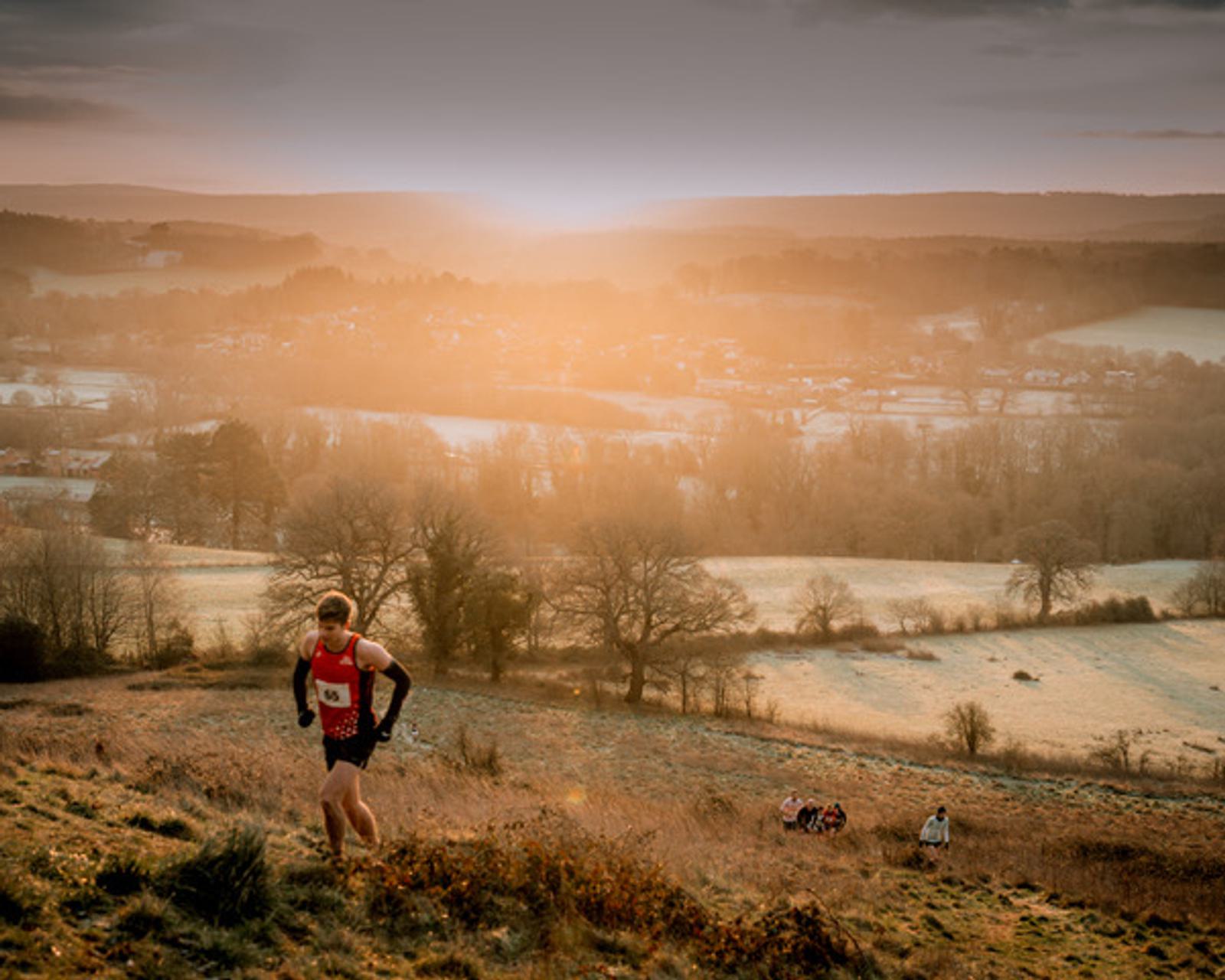 A person wearing a red tank top and black shorts runs up a grassy hill at sunrise. The background features a scenic view of a valley with fields and trees, bathed in warm, golden light.