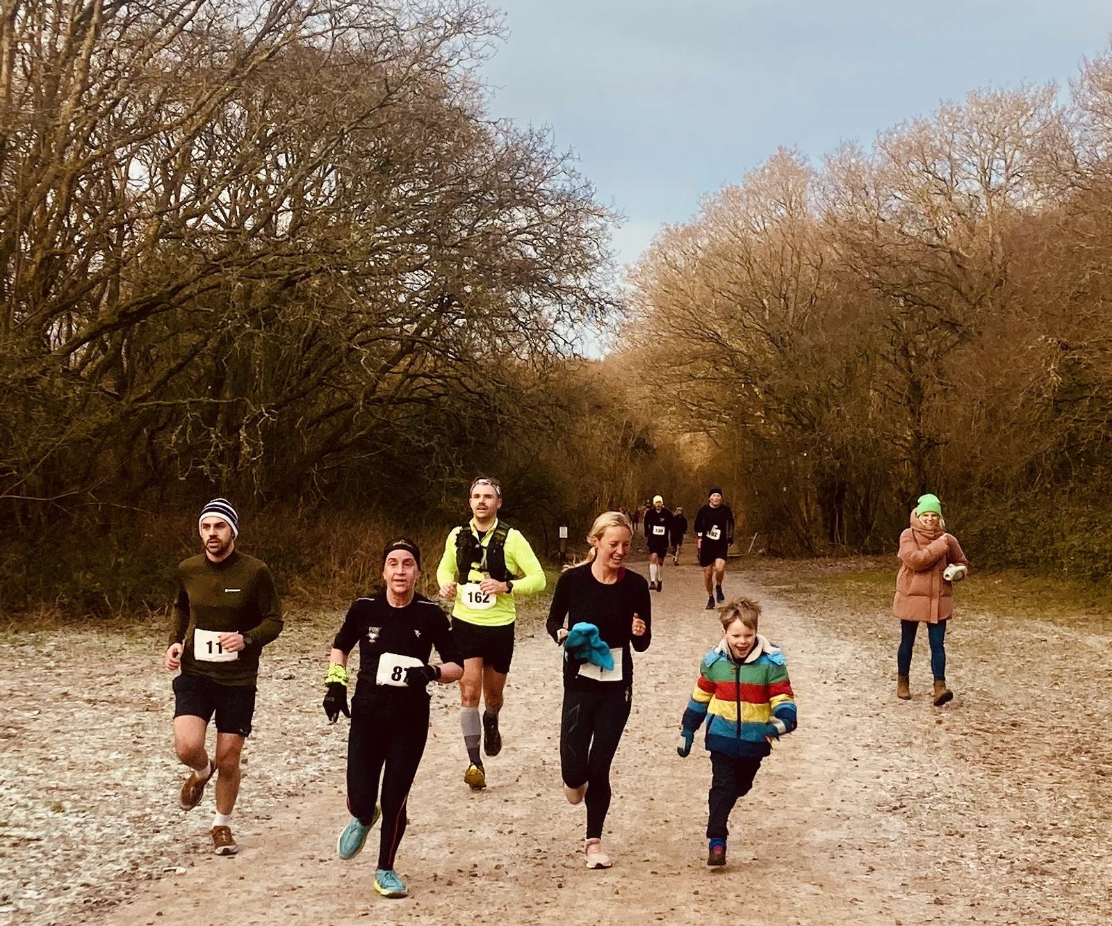 A group of people, including children, are running on a dirt path through a wooded area. They are dressed in athletic gear and appear to be participating in a casual race or jog. Bare trees line the path under an overcast sky.
