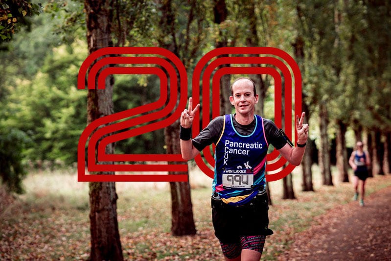 A smiling runner in a Pancreatic Cancer UK shirt holds up both hands in a peace sign while running on a wooded trail, with a large red "20" graphic in the background.