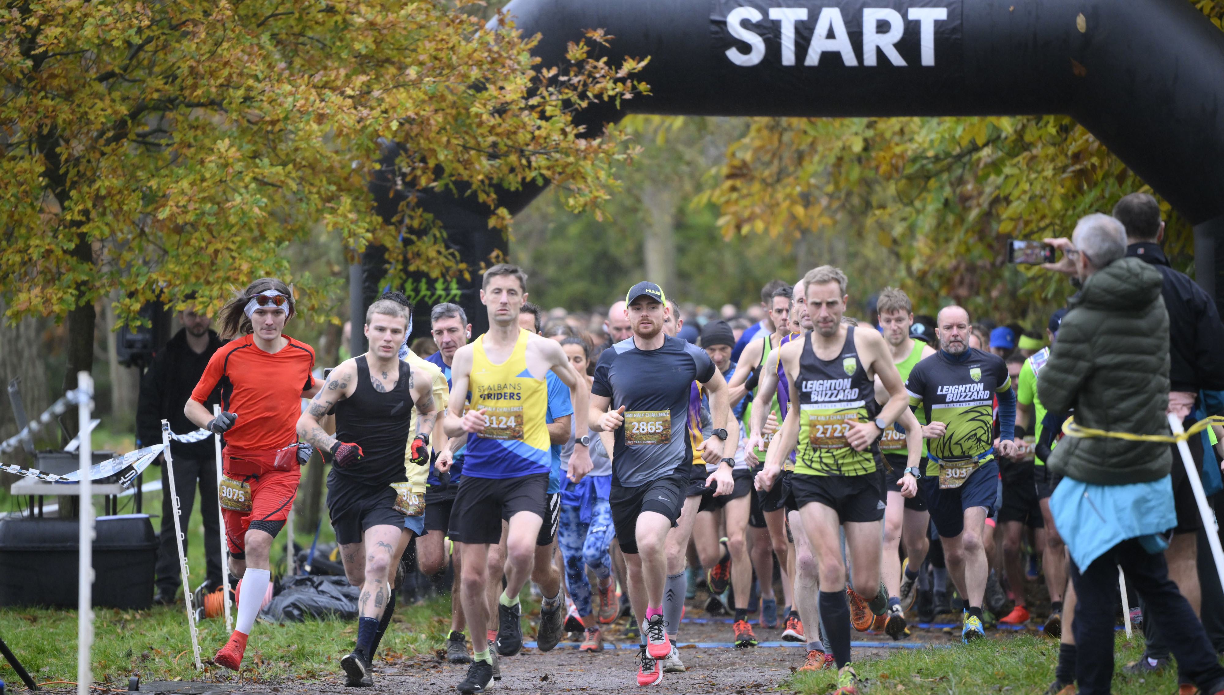 A group of runners in athletic gear start a race under an inflatable arch with "START" written on it. The race takes place in a park with autumn trees and fallen leaves. Spectators and other runners are visible in the background, watching and taking photos.
