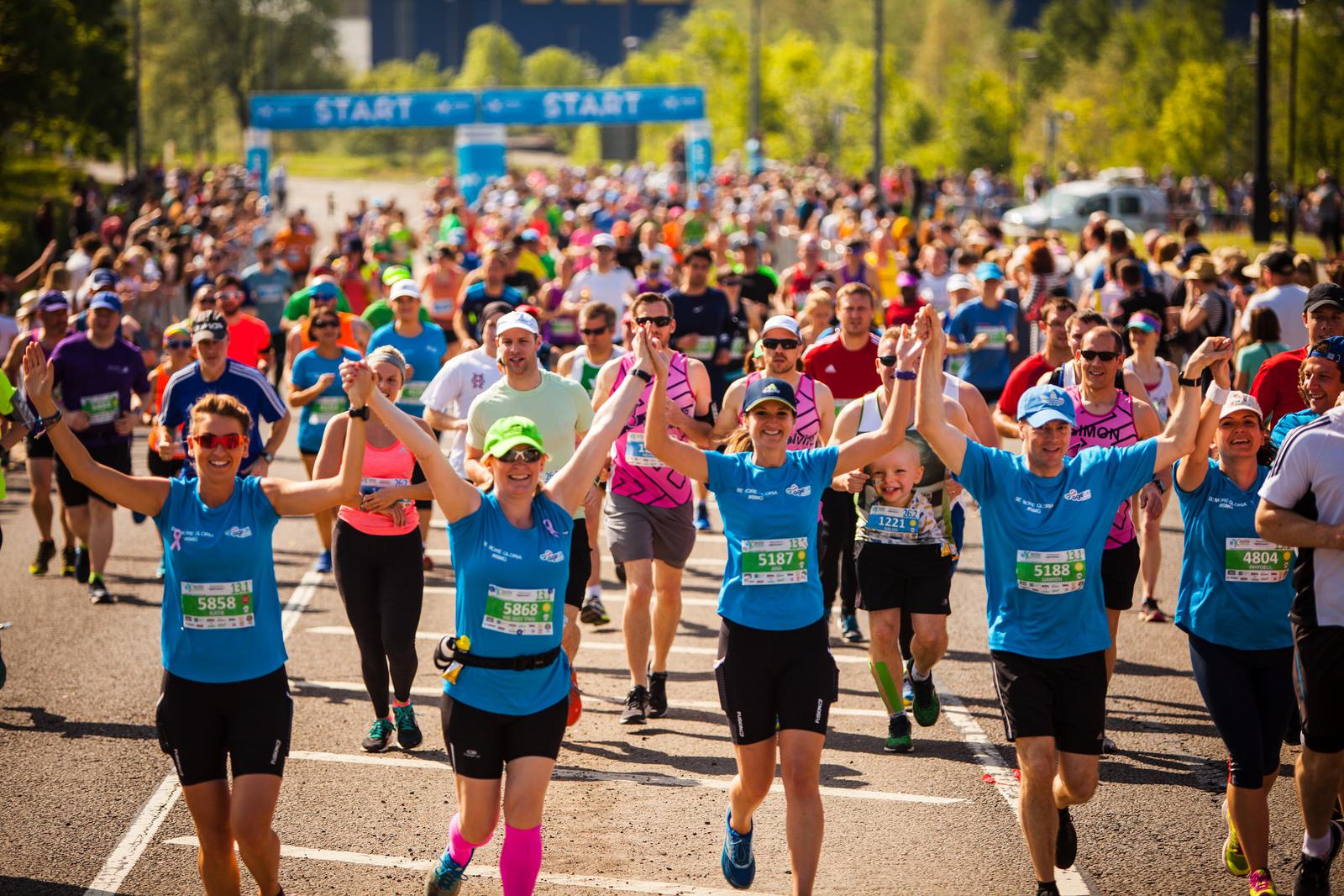 A large group of runners, wearing colorful athletic gear, are participating in a race on a sunny day. The front row features several smiling runners in blue shirts with raised arms, and a "START" banner is visible in the background.
