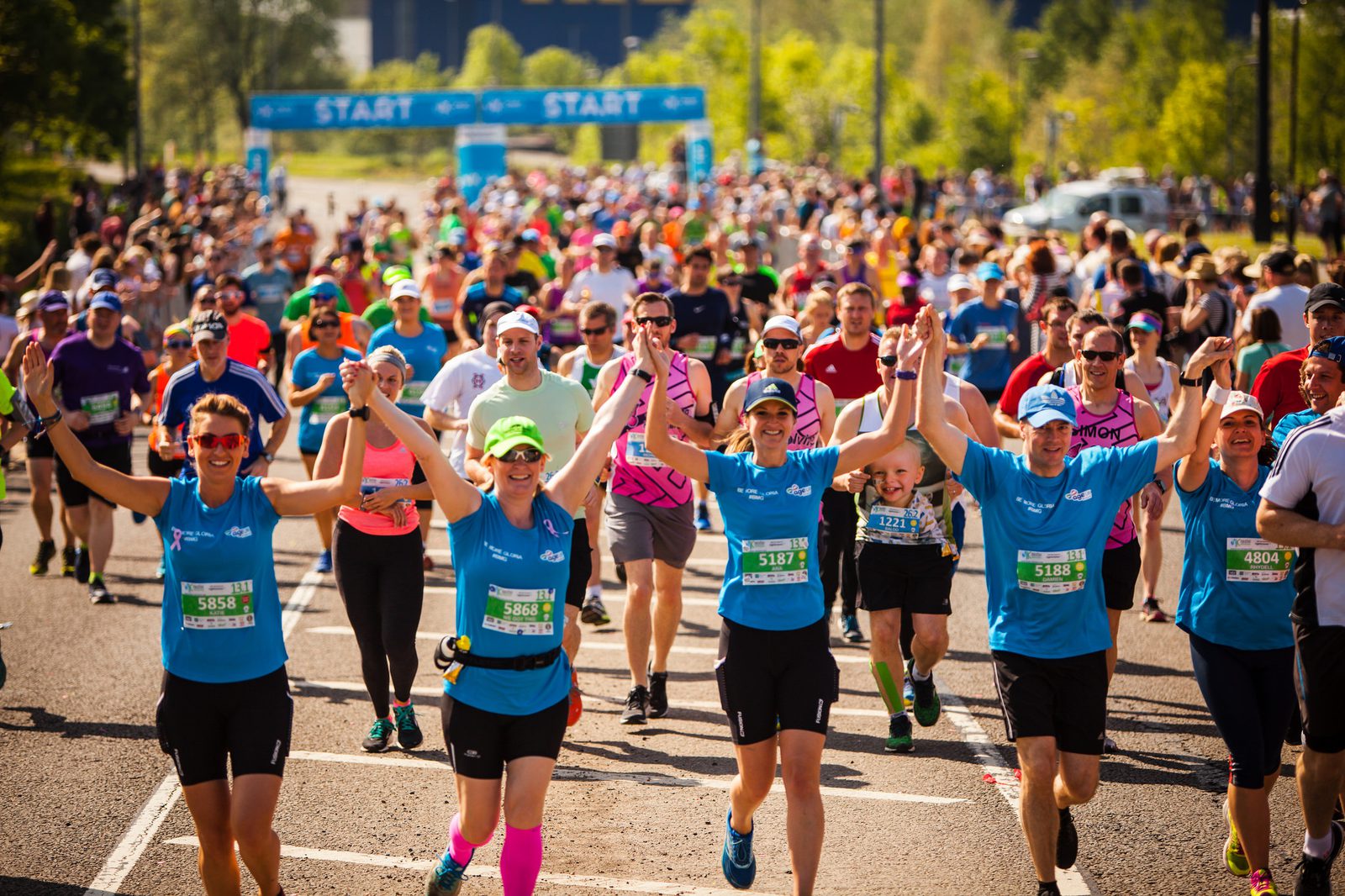 A large group of runners participating in a race, with some at the forefront smiling, holding hands, and raising their arms in triumph. They are running down a road on a sunny day, with the word "START" visible on a banner in the background.