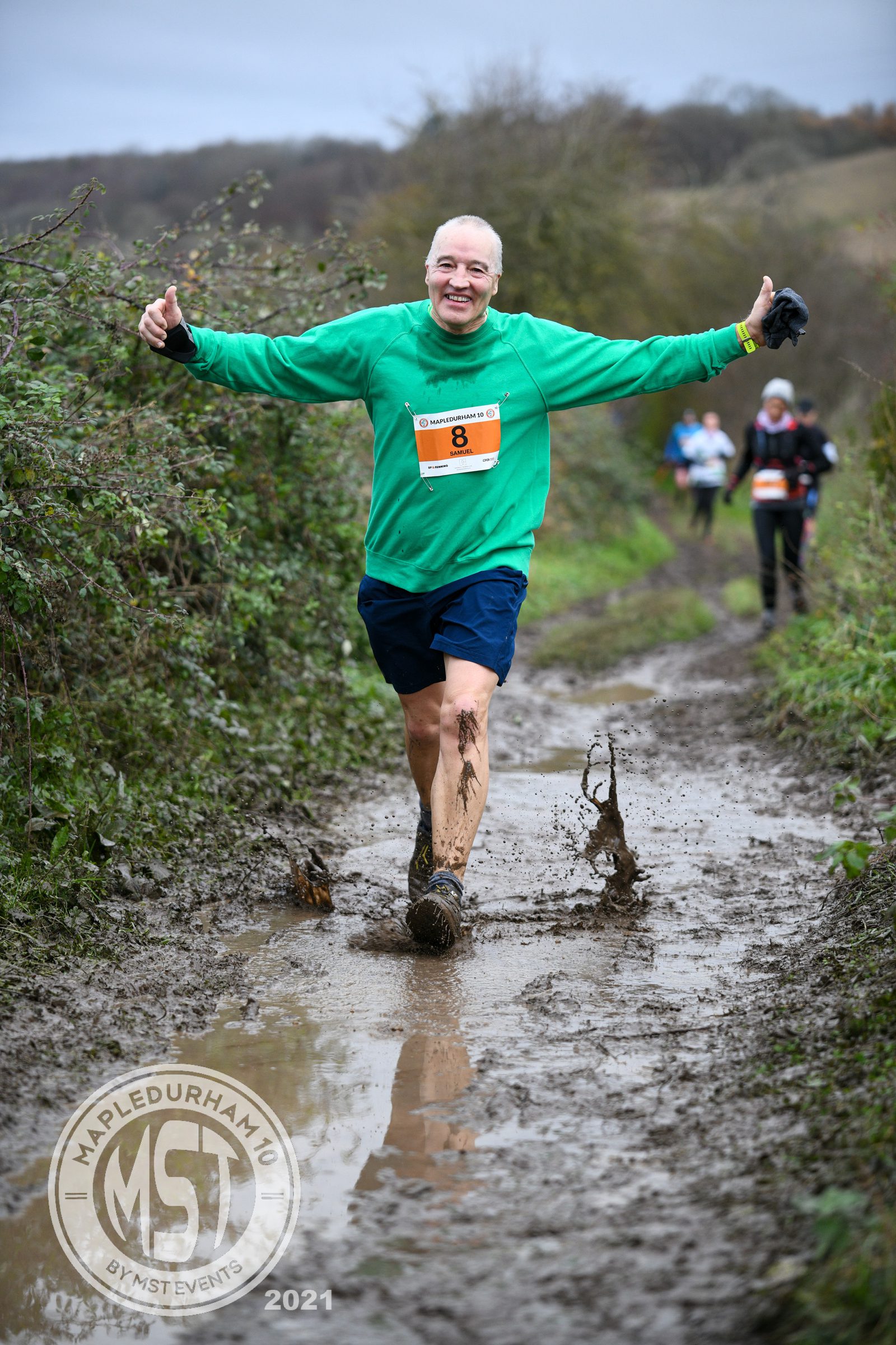 A runner in a green shirt and blue shorts jubilantly splashes through a muddy trail during the Mapledurham 10 event. Mud flies up from his shoes as he stretches his arms out, projecting enthusiasm. Other runners can be seen further down the trail. The event logo and year 2021 are visible.