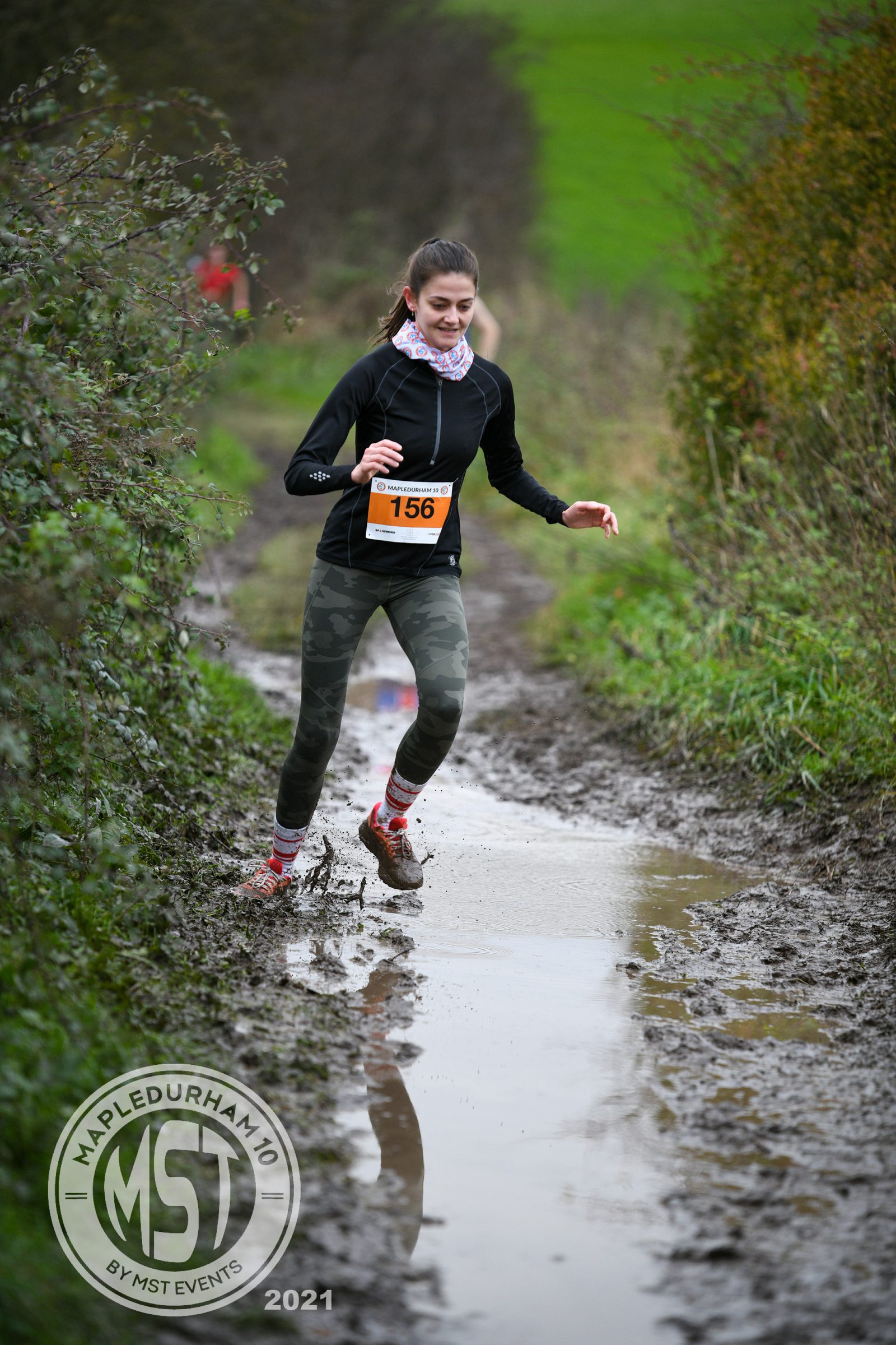 A woman wearing a black jacket, camouflage leggings, and a bib numbered 156 is running on a muddy trail. She is mid-step, splashing through a puddle. The surrounding area is wooded and green, with the MST logo and "Mapledurham" watermark in the bottom left corner.