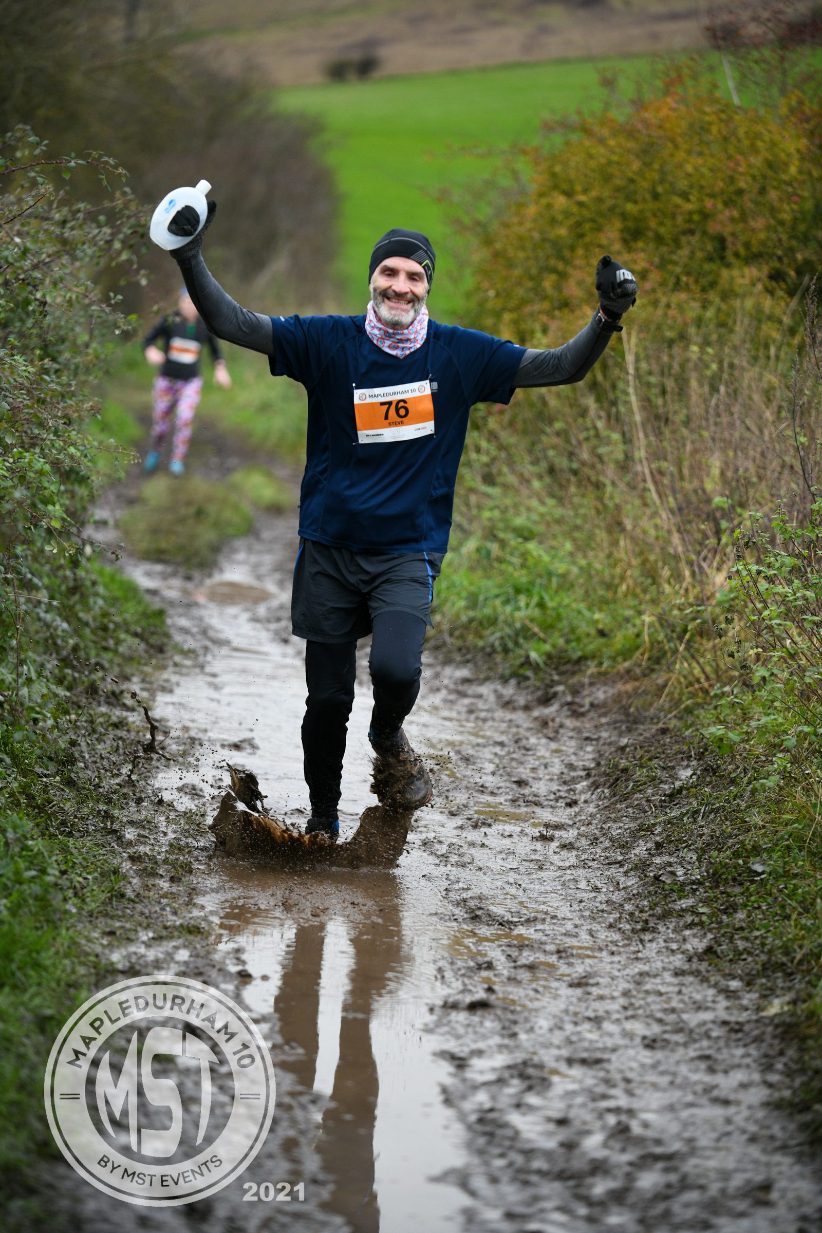A runner wearing a black outfit and beanie, with bib number 76, triumphantly raises both arms while running through a muddy trail in an outdoor race. Visible mud splashes on his legs and ground. The event logo in the corner reads, "Mapledurham 10 by MST Events 2021.