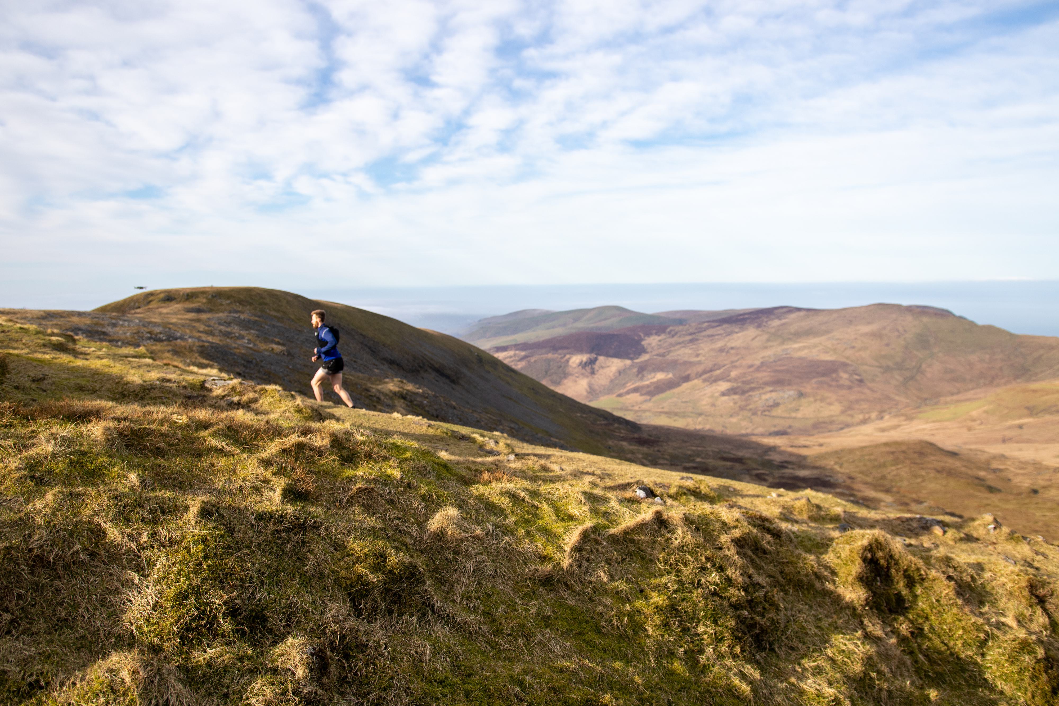 A person in athletic wear is running uphill on a grassy mountainside under a partly cloudy sky. The landscape features rolling hills and distant mountains, suggesting a remote and scenic outdoor location.