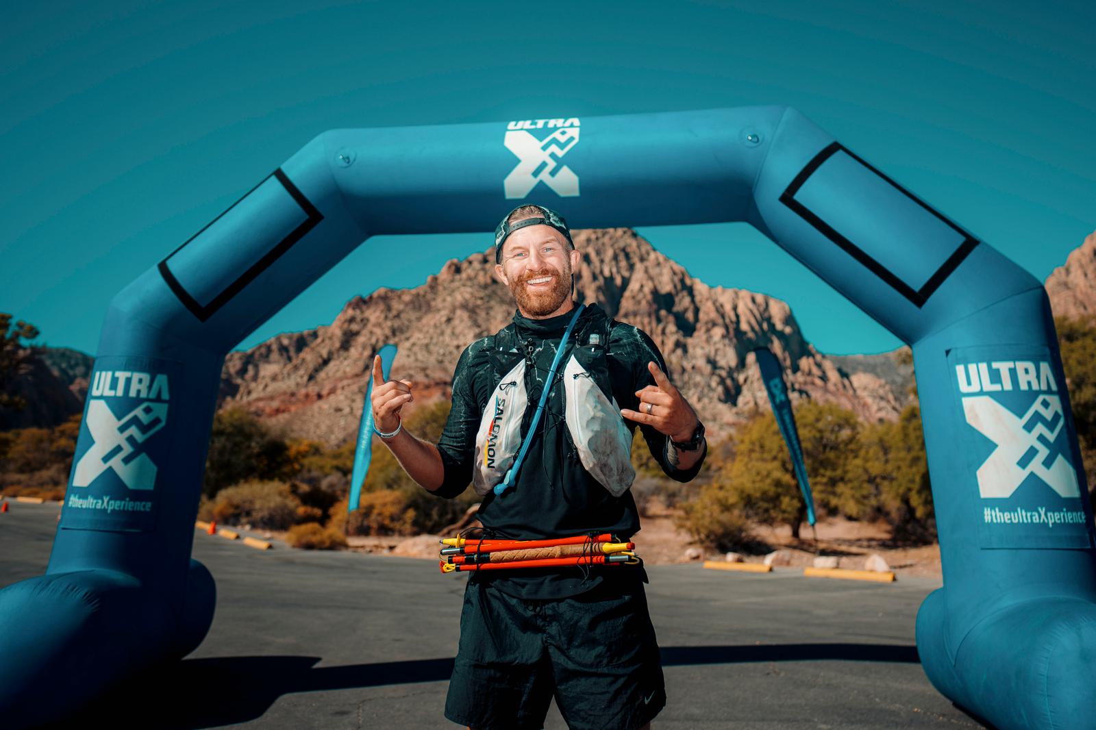 A person in athletic gear smiles and gestures with their hands at the finish line of a race, standing under a blue inflatable arch that reads "ULTRA X." In the background, there are rocky hills and clear blue skies.