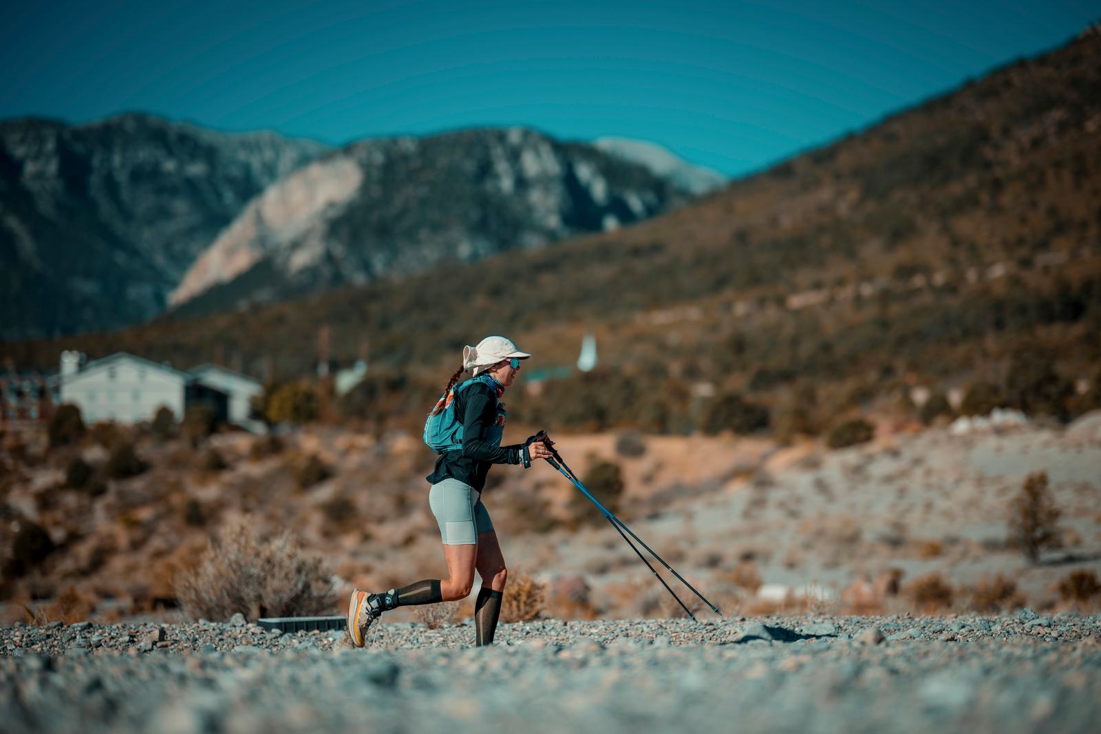 A person with a prosthetic leg is hiking on a rocky path using trekking poles. They are wearing a cap and carrying a backpack. The background features mountains and a clear blue sky.