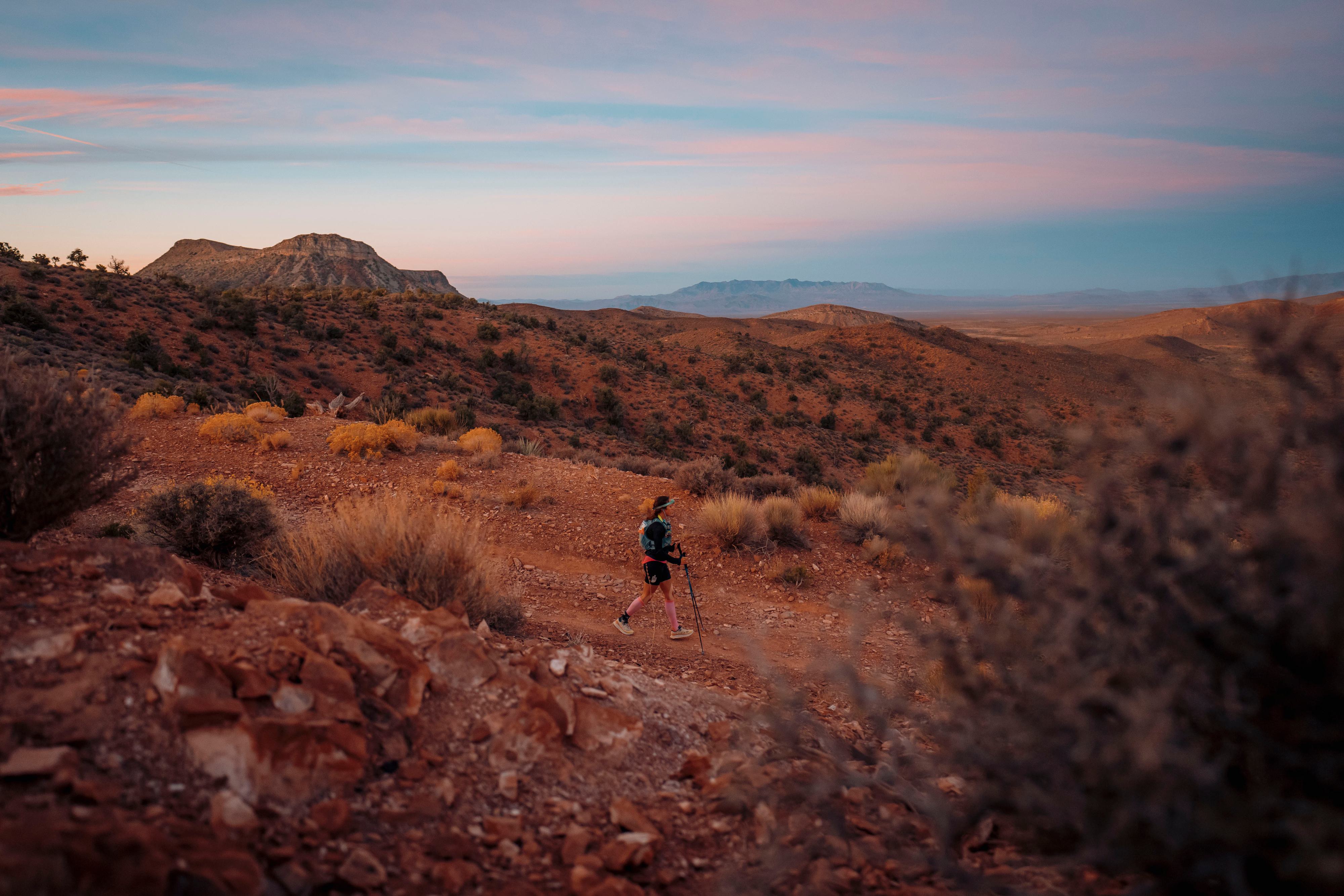 A person hikes along a rocky desert trail at sunset, surrounded by scrubby vegetation and distant hills. The sky is painted with soft pink and blue hues, creating a serene and expansive landscape.