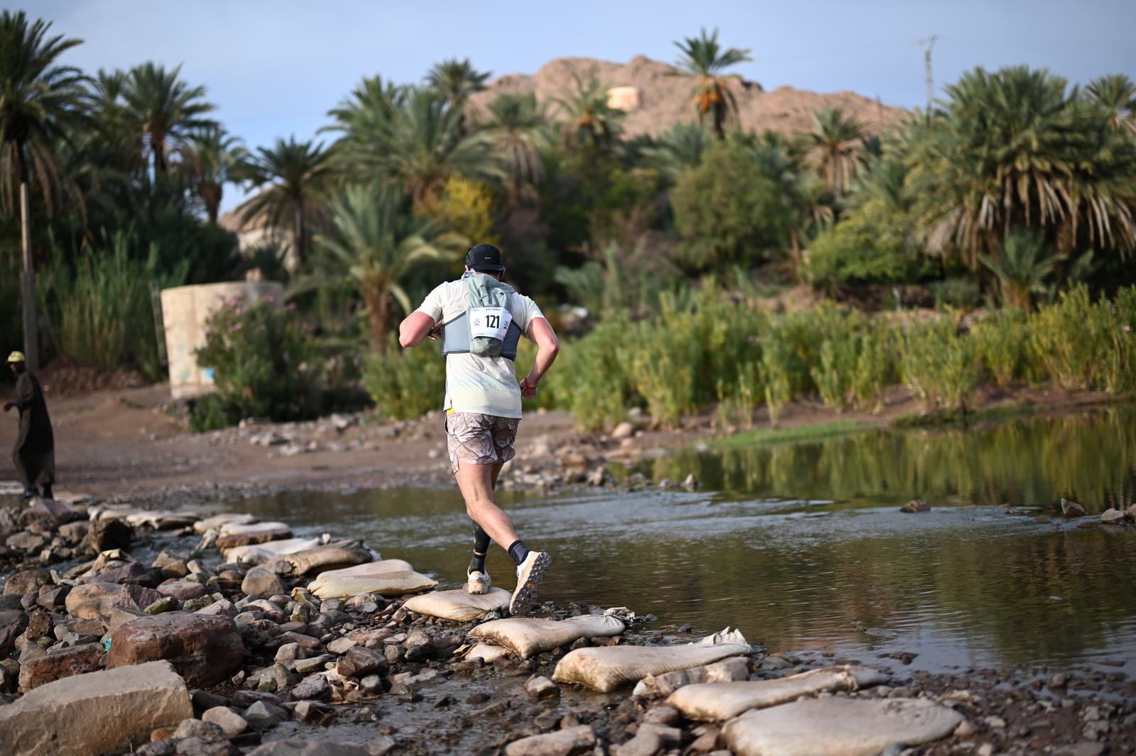 A runner wearing a numbered vest is crossing a rocky stream using stepping stones. The background features lush greenery, palm trees, and a rocky hillside under a clear sky.