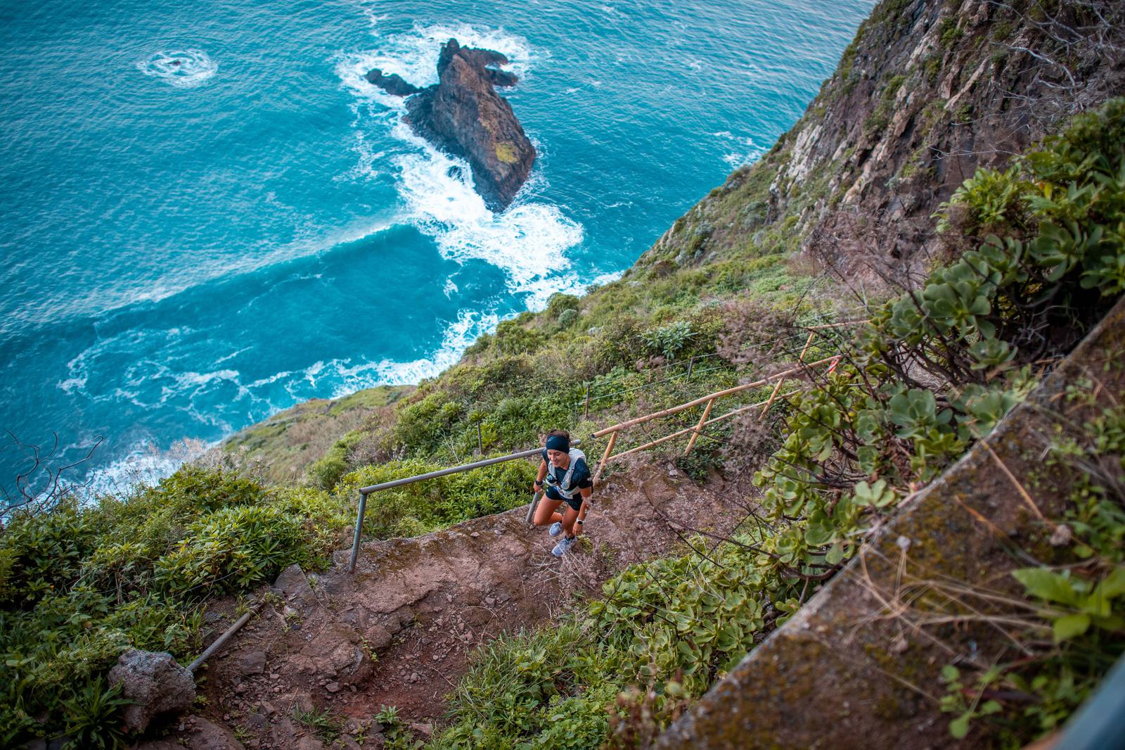 A person is hiking up a steep, narrow trail with a metal railing along a rocky, green-clad cliffside. Below, the ocean is vibrant blue, with waves crashing against a large rock. The view is expansive and dramatic.