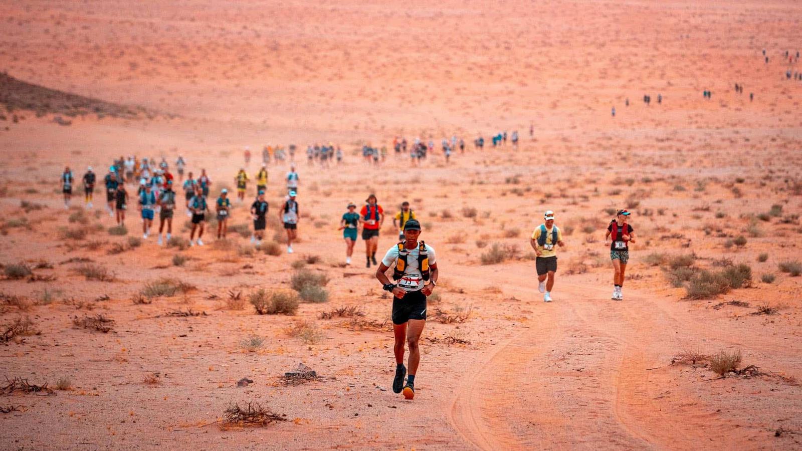 A group of runners spread out over a vast, sandy desert landscape. The lead runner is in focus, wearing a number bib and athletic gear. The sky is clear, and small shrubs dot the reddish terrain underfoot.