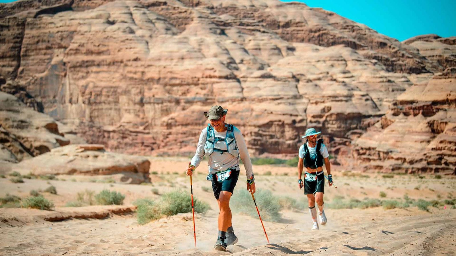 Two people hike through a desert landscape with rocky cliffs in the background. Both wear hats, backpacks, and use trekking poles. The sky is clear and blue.