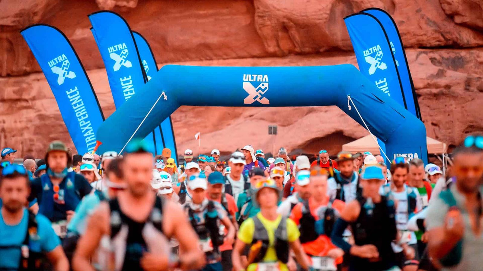 A large group of runners participate in an outdoor marathon event, passing under a blue inflatable arch with "Ultra X" written on it. They are surrounded by rocky, desert-like terrain.