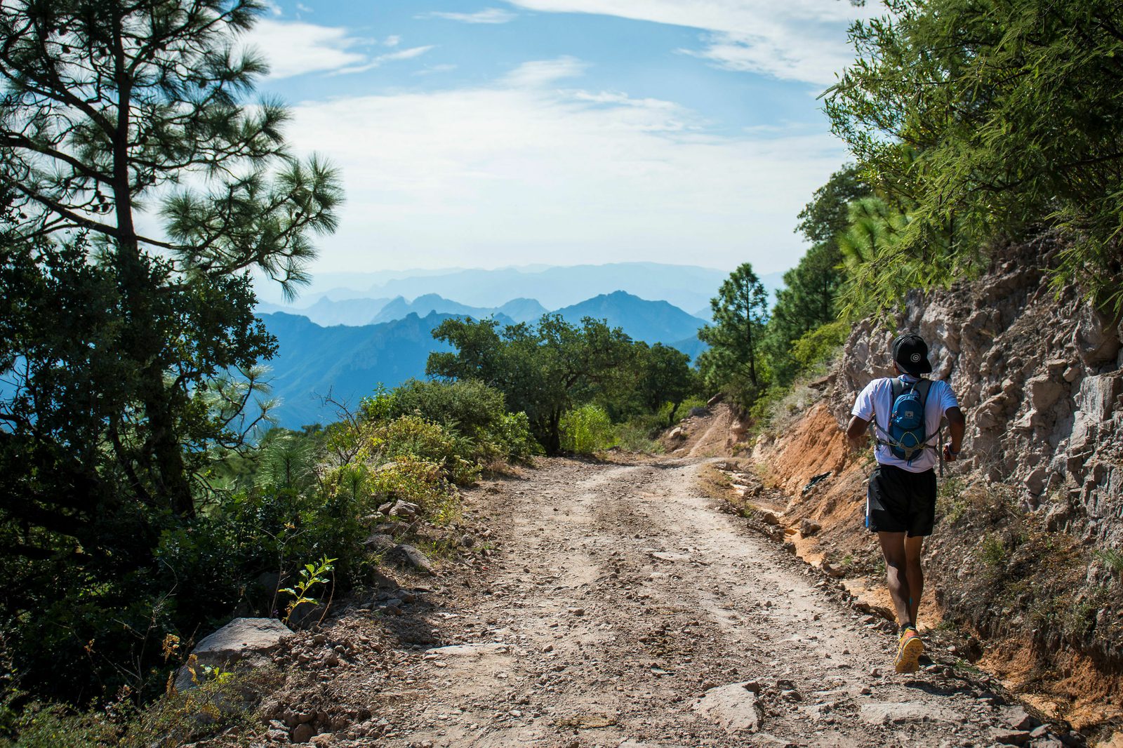 A person is running on a rocky, dirt trail surrounded by lush greenery and trees, with a mountainous landscape in the background. The sky is clear with a few scattered clouds, and the scene exudes a sense of adventure and tranquility.