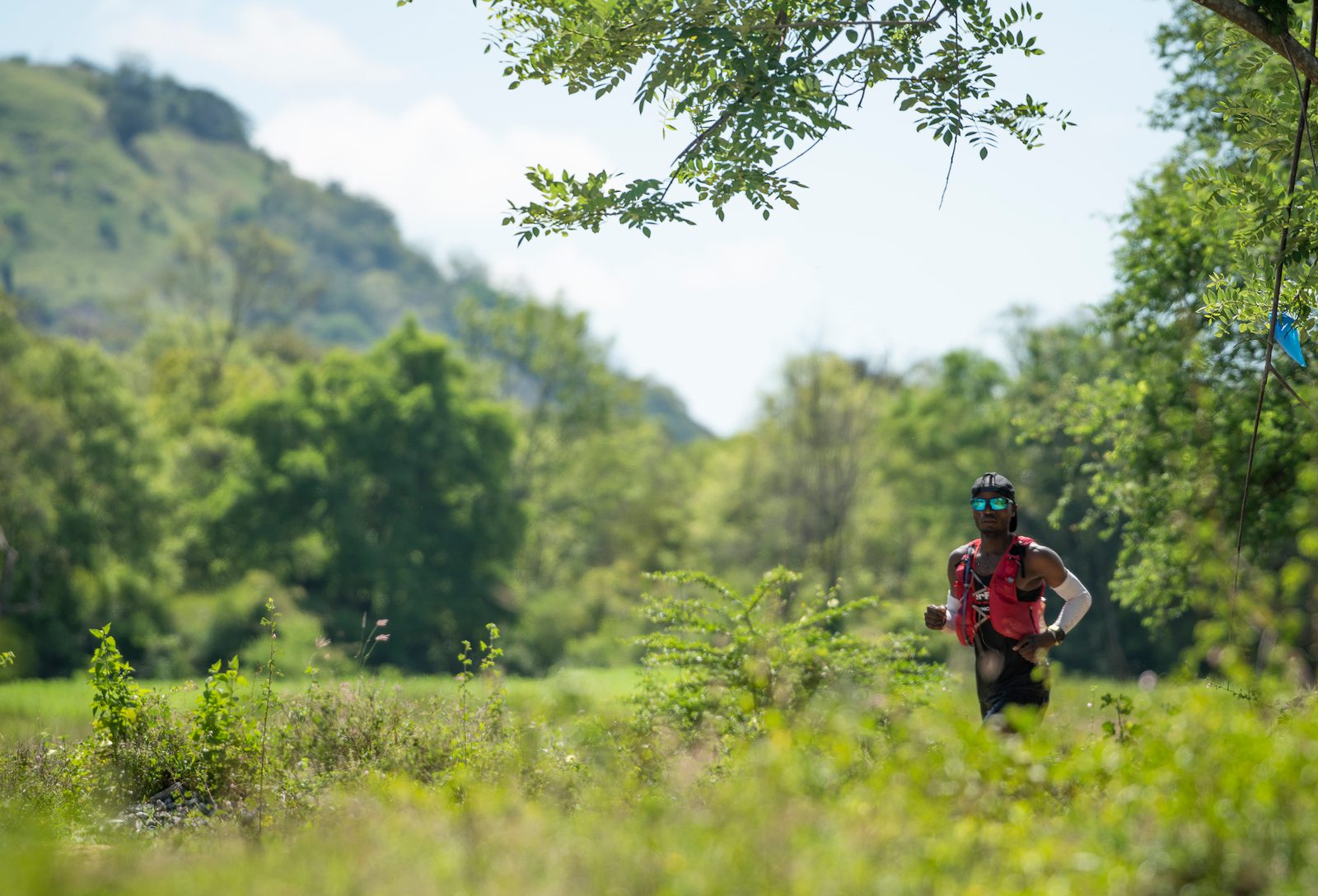 A person wearing a red vest and dark clothing is running through a lush, green field. They are surrounded by trees and vegetation with hills visible in the background under a clear sky.