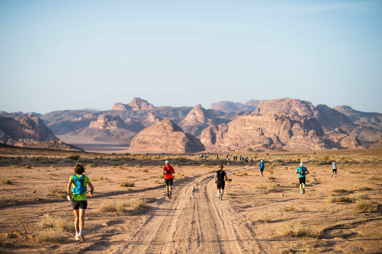 Runners are participating in a race across a vast desert landscape with rocky mountains in the background. The ground is sandy and dotted with sparse vegetation, and the sky is clear, suggesting a sunny day. The runners wear athletic gear and backpacks.