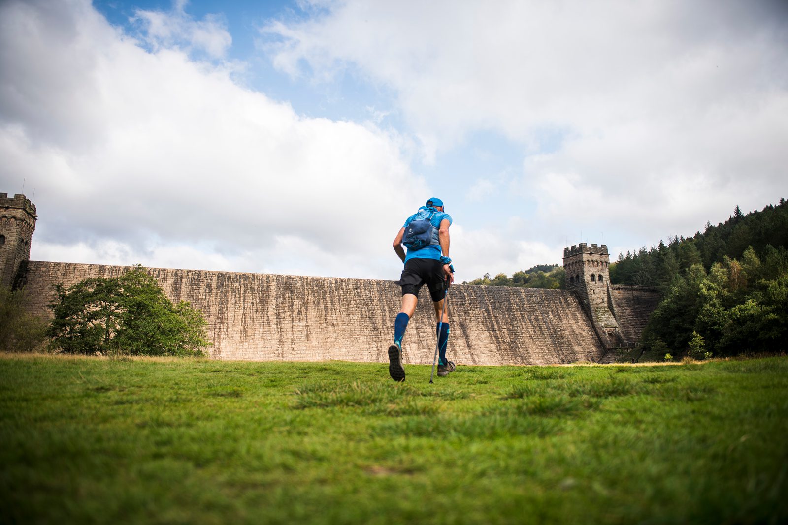 A person wearing a blue outfit and carrying a backpack runs across a grassy field towards a large stone dam, with two towers on either side. The sky above is partly cloudy. Trees can be seen on the right side of the dam.
