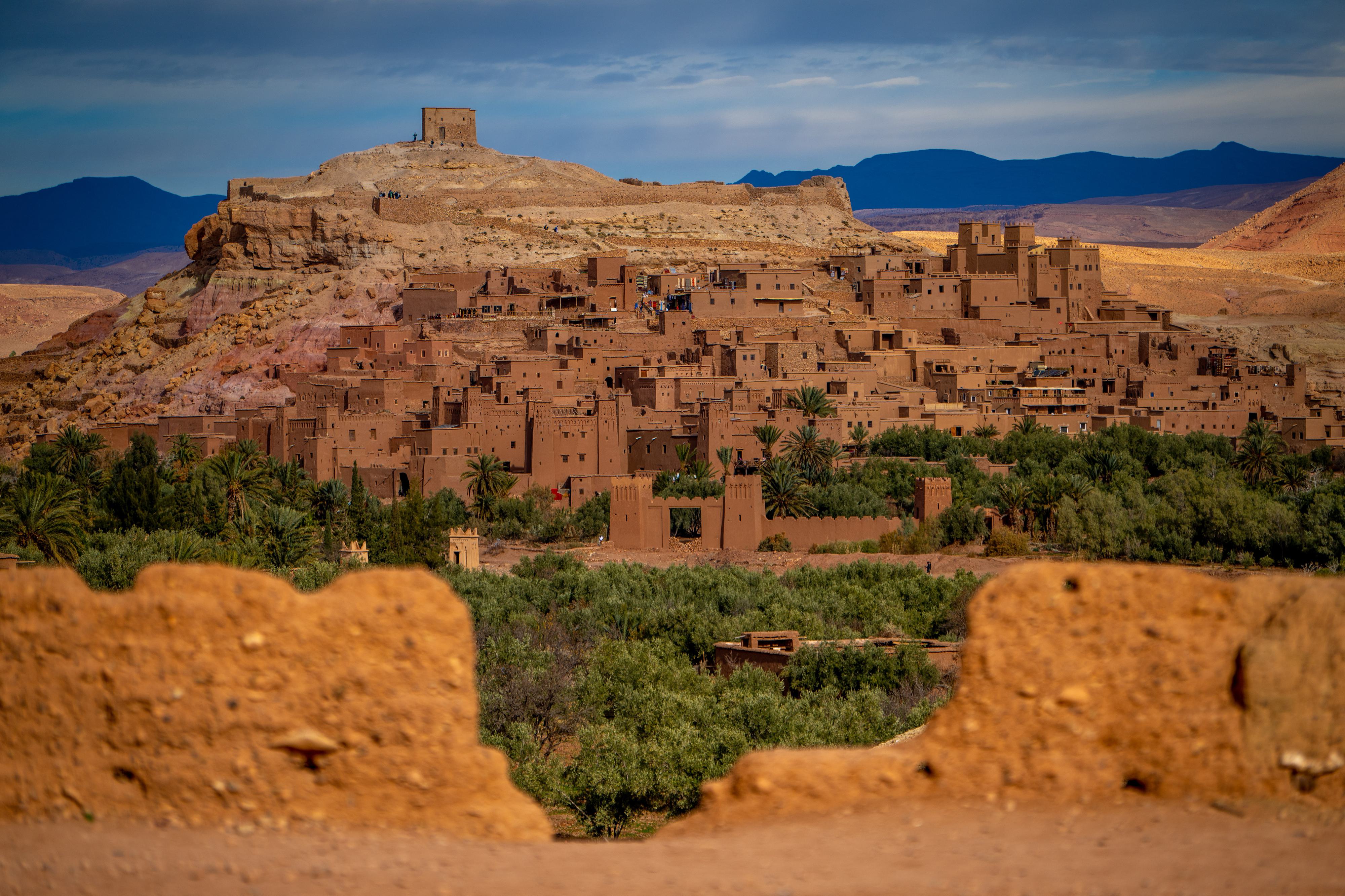 A scenic view of Ait Benhaddou, an ancient fortified village in Morocco. Clay and mud-brick buildings are stacked on a hillside, surrounded by a lush green landscape and mountains in the background under a blue sky.