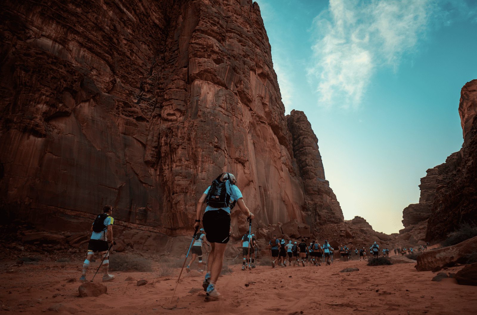 A group of people wearing athletic gear hike through a narrow canyon with towering, rugged red rock cliffs under a clear blue sky. The scene suggests an organized trek or race in a desert landscape.
