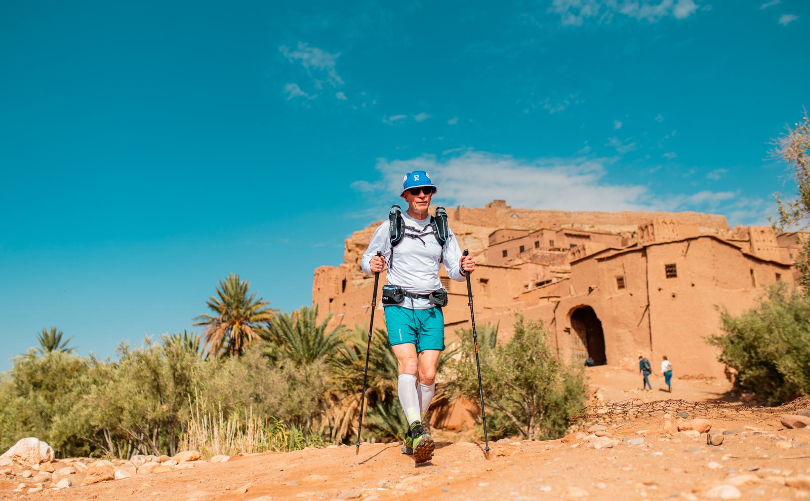 A person in outdoor gear and a hat is hiking with poles on a rocky path near an ancient clay building and palm trees, under a bright blue sky. Another person is visible in the background.