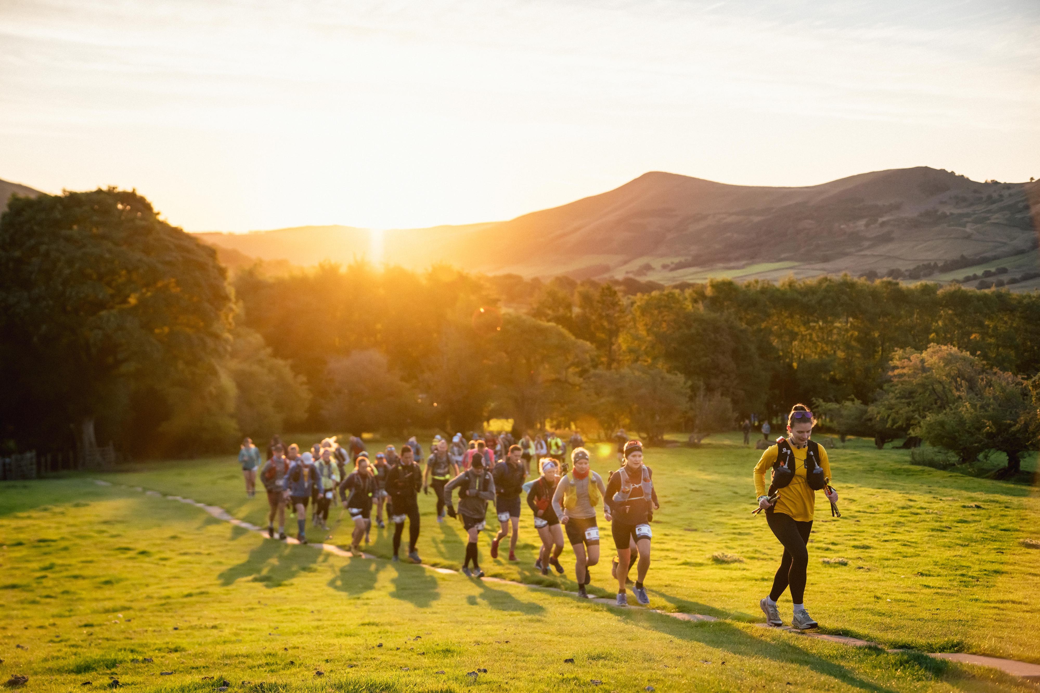 A group of people are hiking on a grassy path through a scenic landscape at sunset. The path winds through lush green hills and trees with mountains in the background. The sun is setting, casting a warm golden glow over the group and the surroundings.