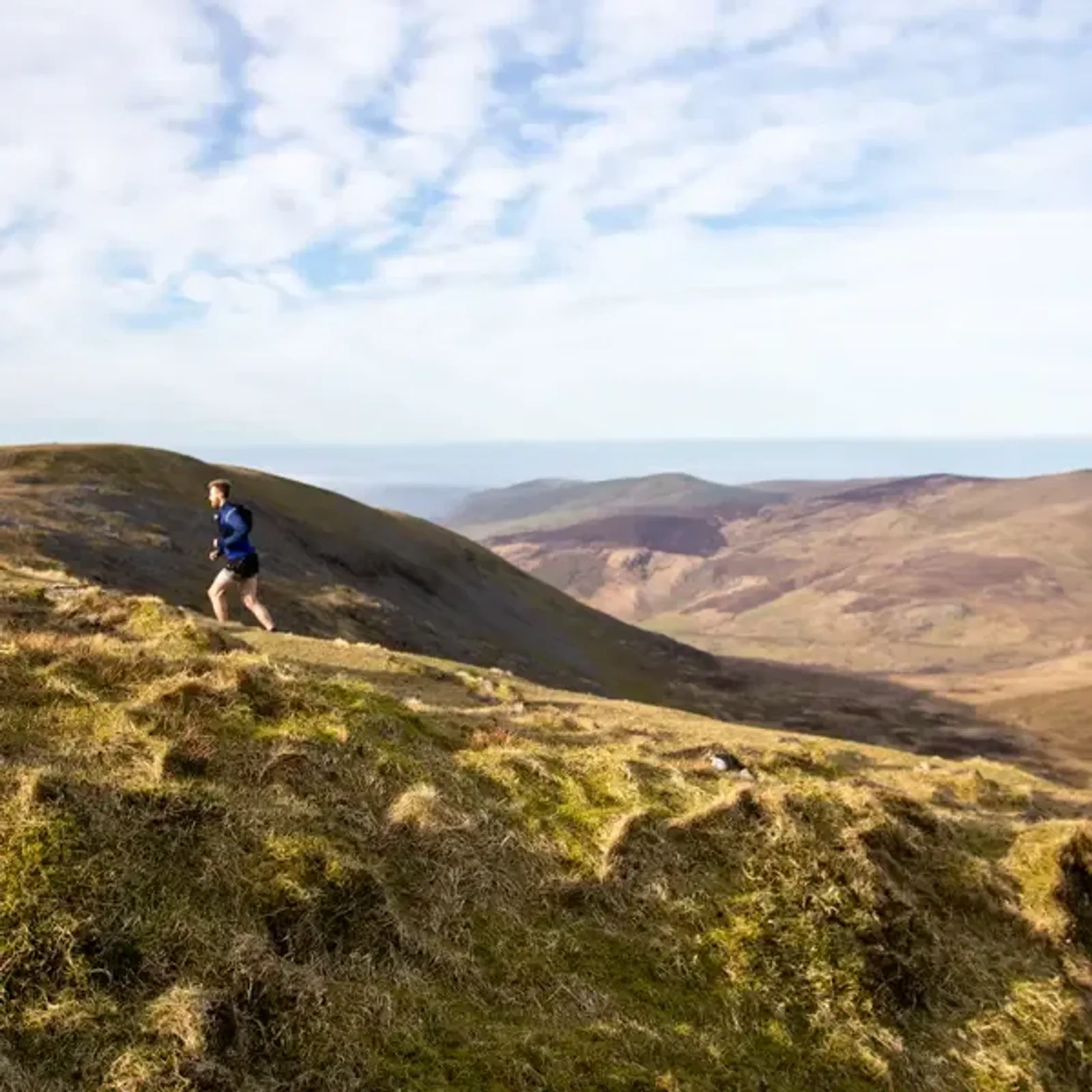 A person is running along a grassy ridge in a mountainous landscape under a partly cloudy sky. The scene features rolling hills and distant peaks, with the runner dressed in a blue shirt and shorts.
