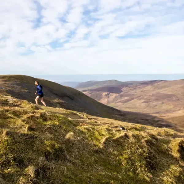 A person is running along a grassy ridge in a mountainous landscape under a partly cloudy sky. The scene features rolling hills and distant peaks, with the runner dressed in a blue shirt and shorts.