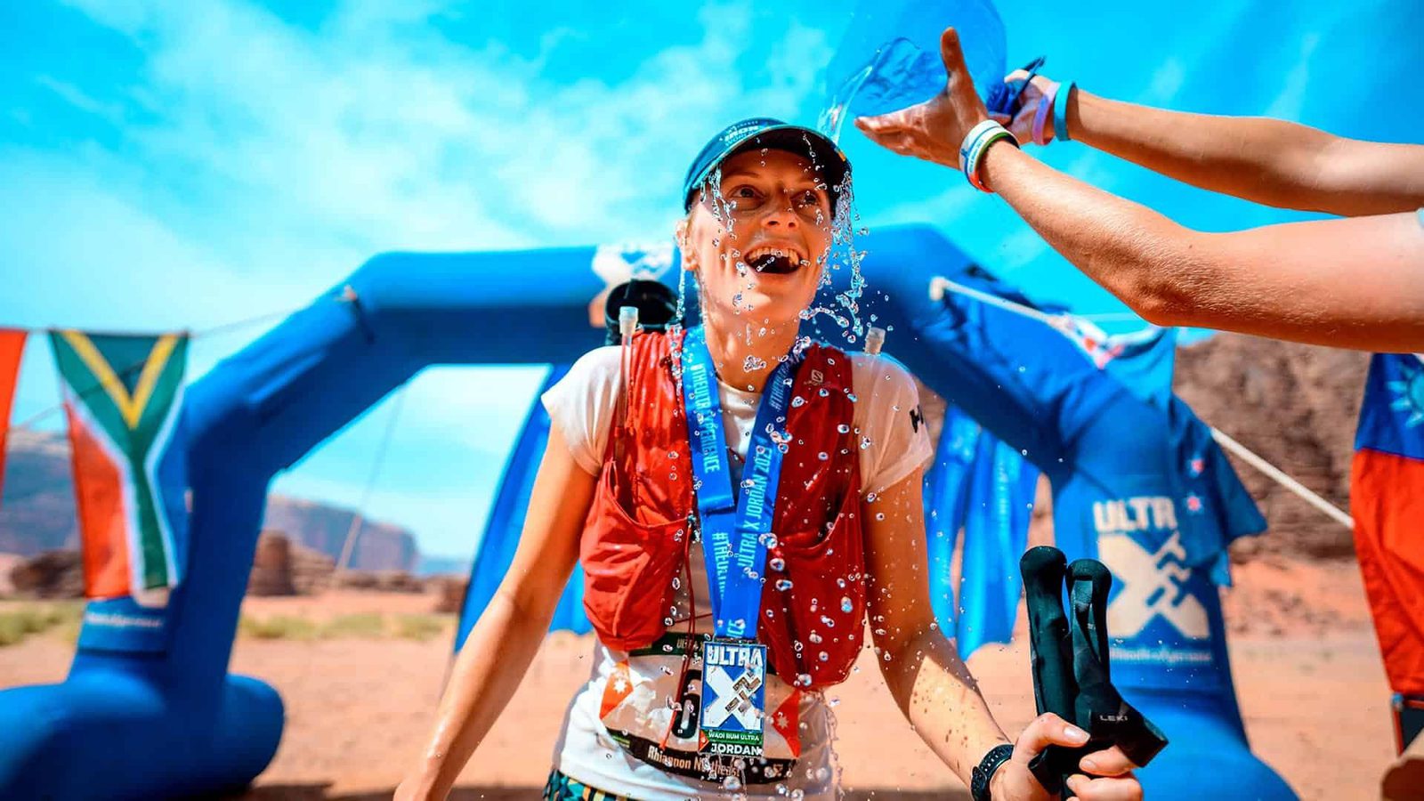 A joyful runner wearing a medal stands at a race finish line, being splashed with water under a clear blue sky. The scene is surrounded by flags and an inflatable arch in a desert landscape.