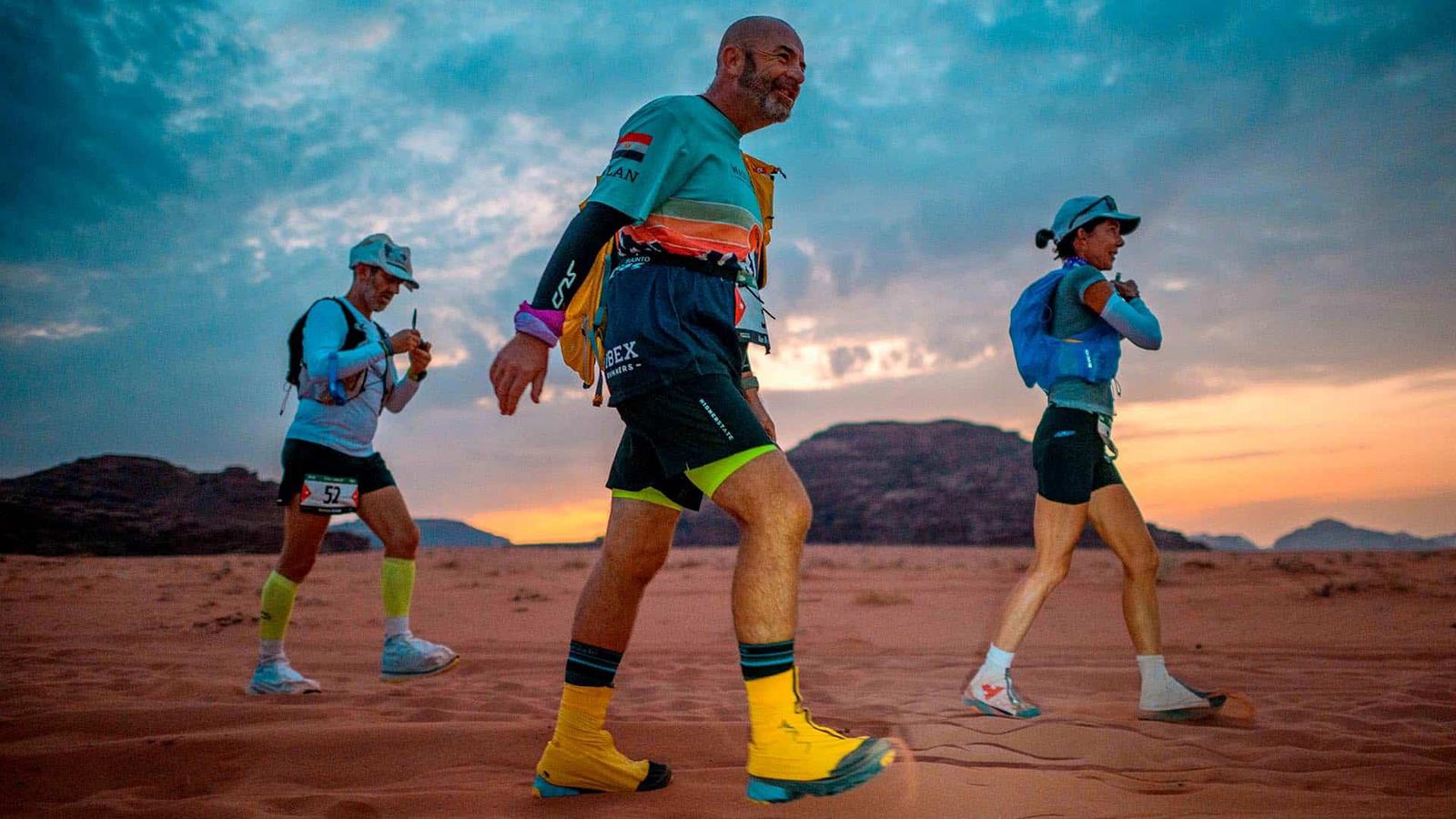Three runners in vibrant gear traverse a desert landscape at dusk. The sky is a mix of blues and oranges as the sun sets behind distant rocky hills. They carry backpacks and wear caps, moving energetically across the sandy terrain.