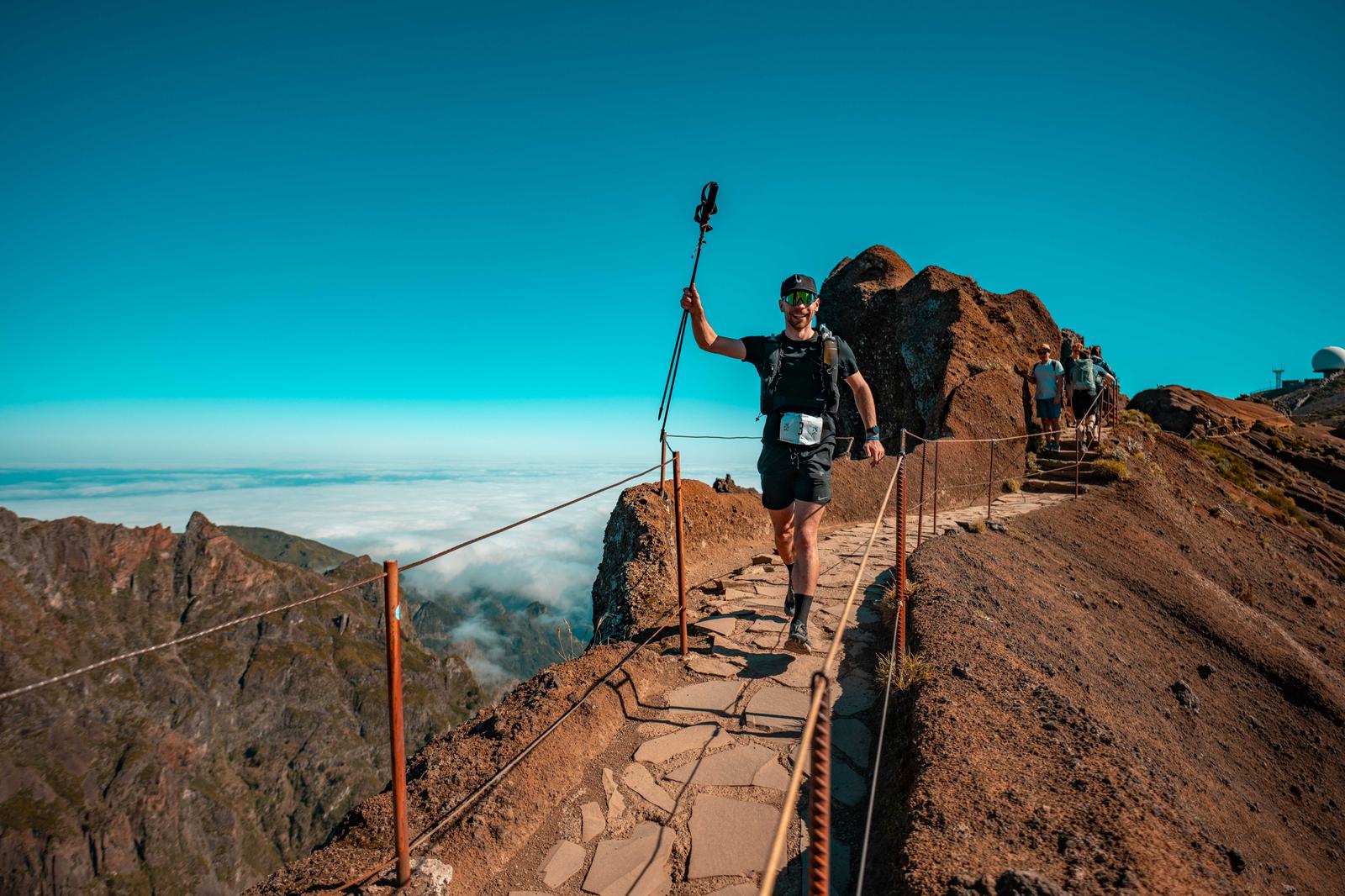 A person in athletic gear smiles while hiking on a narrow mountain path under a clear blue sky. They hold trekking poles and are surrounded by stunning rocky landscapes and distant mountains. Other hikers are visible in the background.