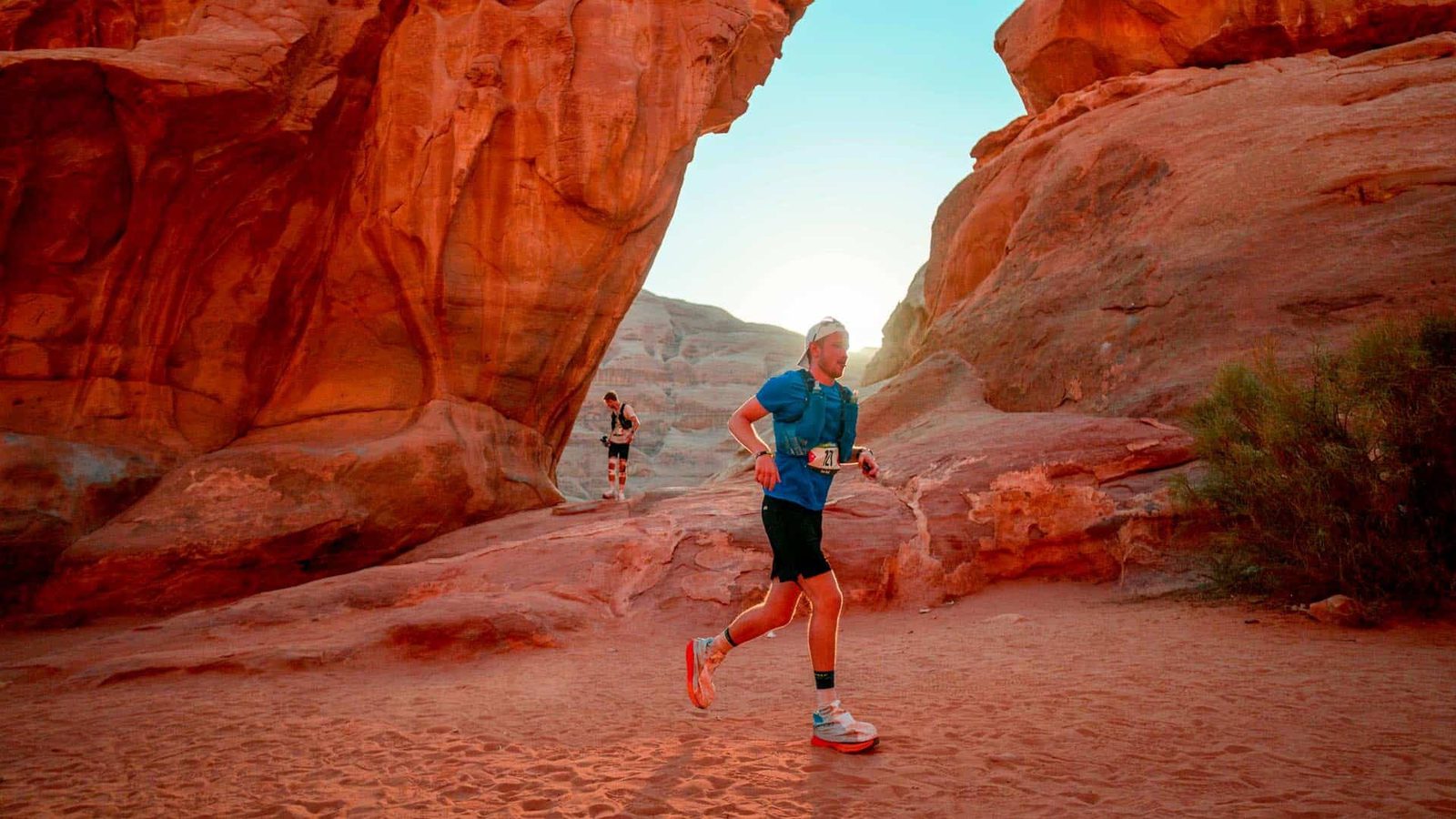 Two runners jog through a desert landscape with large red rock formations. The sky is clear and blue, and the sun is low in the background, casting warm light on the scene. The foreground runner wears a blue shirt and black shorts.