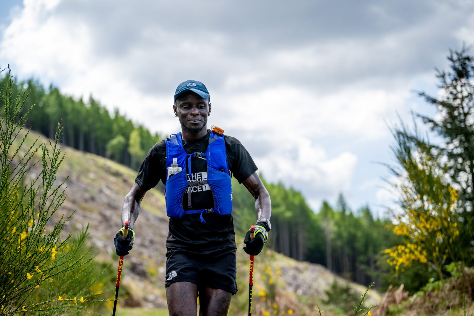 A person wearing a blue hydration vest and black athletic outfit runs with trekking poles on a trail in a lush, mountainous area under a cloudy sky. Green bushes with yellow flowers border the path, and tall trees are visible in the background.