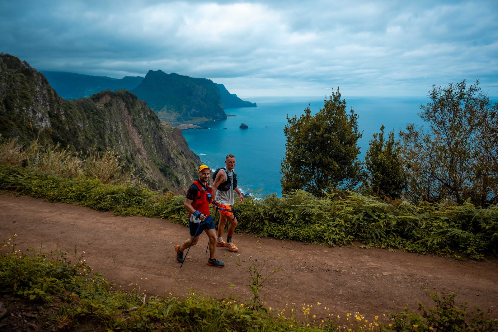 Two people are hiking on a dirt trail along a lush coastline. The ocean and rugged cliffs form a dramatic backdrop under a cloudy sky. One hiker wears a red top and the other a gray top.