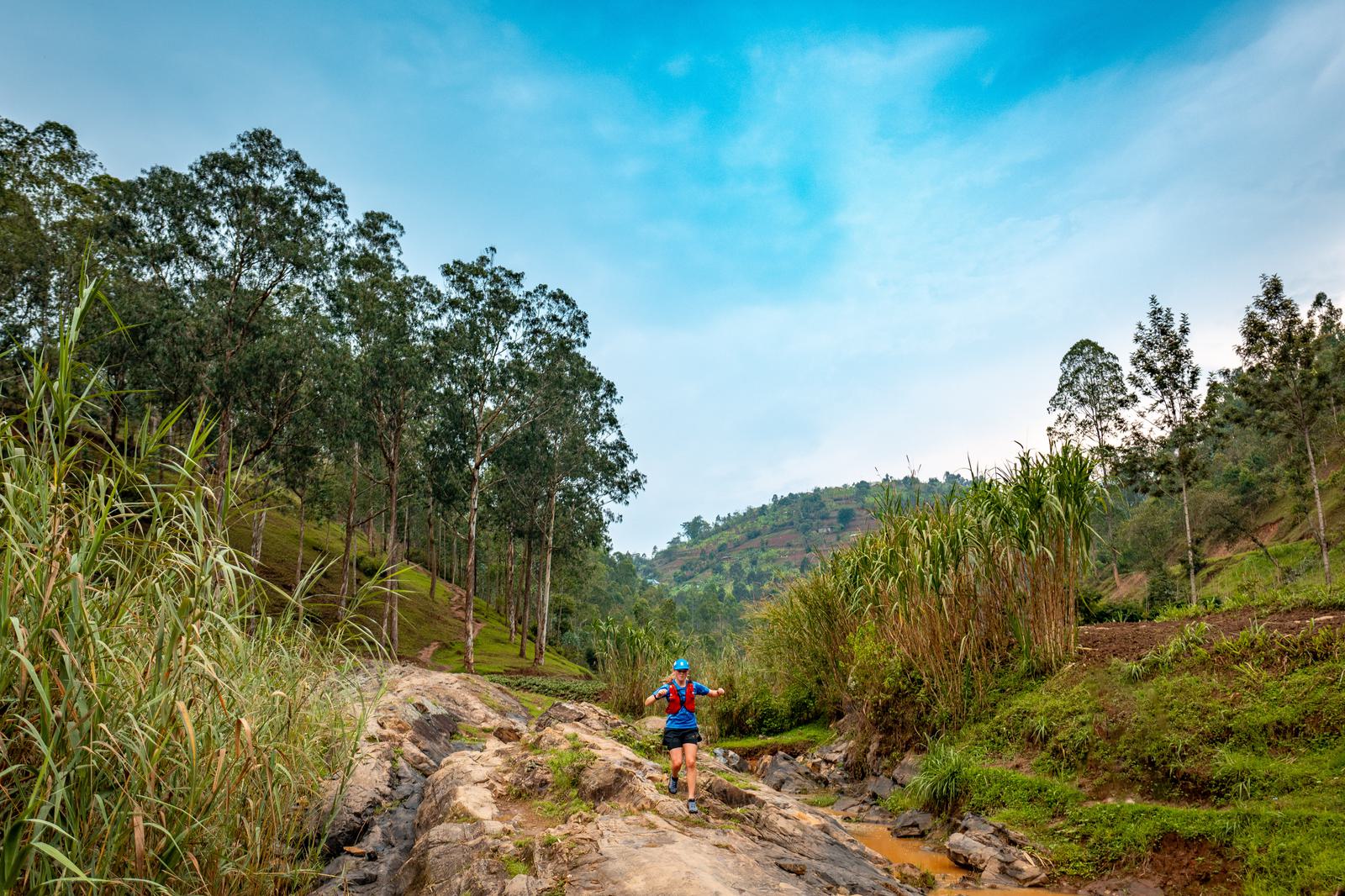A person wearing a blue helmet and red shirt is hiking on rocky terrain surrounded by tall grasses and trees. The sky is clear with a few clouds, and a hill covered in green foliage is visible in the background.
