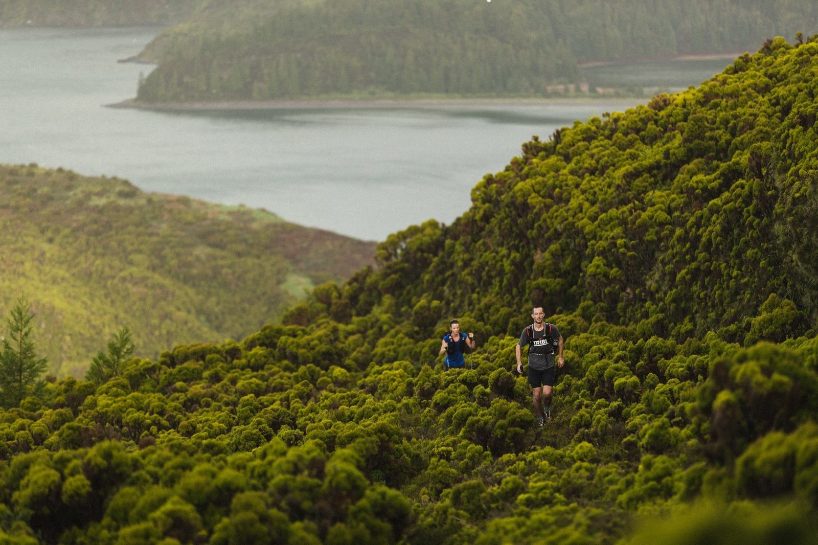 Two people running along a narrow, rugged trail surrounded by lush, green vegetation on a hillside. A body of water and more forested hills are visible in the background under overcast skies. The scene exudes a sense of adventure and natural beauty.