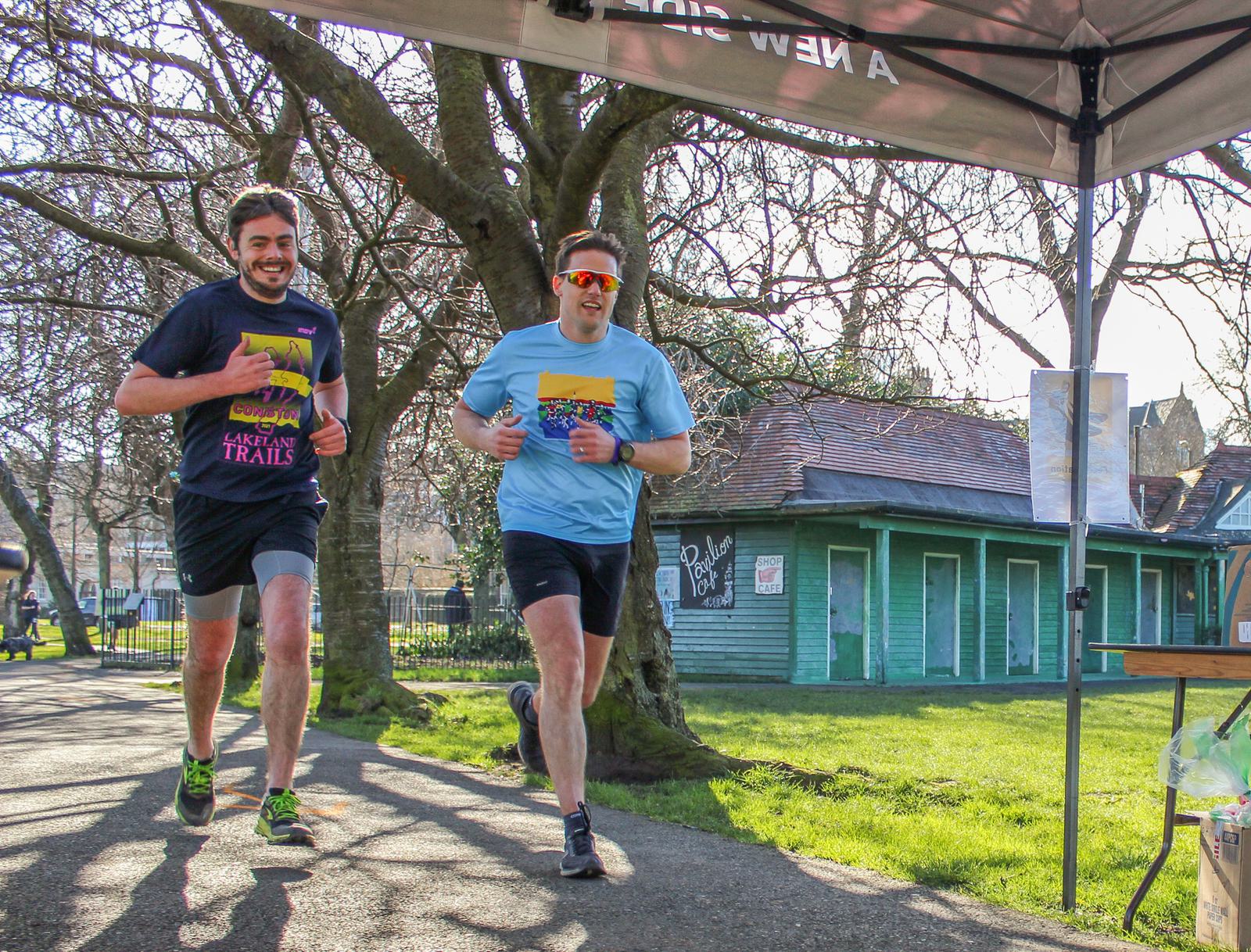 Two men are running side by side on a park pathway during a race. Both are wearing colorful shirts and sunglasses. Trees and a green building with graffiti are in the background. A pavilion canopy is partially visible overhead. The weather is sunny.